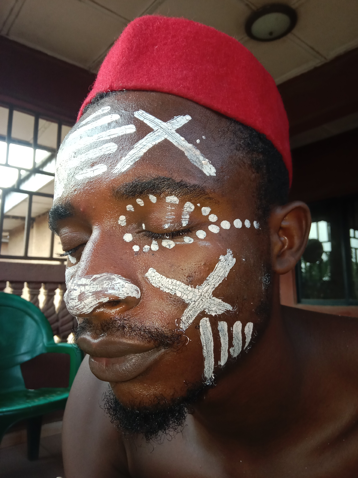 A man with dark skin and a beard wears a red hat and white paint markings on his face. The paint markings include lines, crosses, and dots. He has his eyes closed. He is in an outdoor setting, possibly a porch, with a green plastic chair visible in the background. The location is Unknown, Unknown.