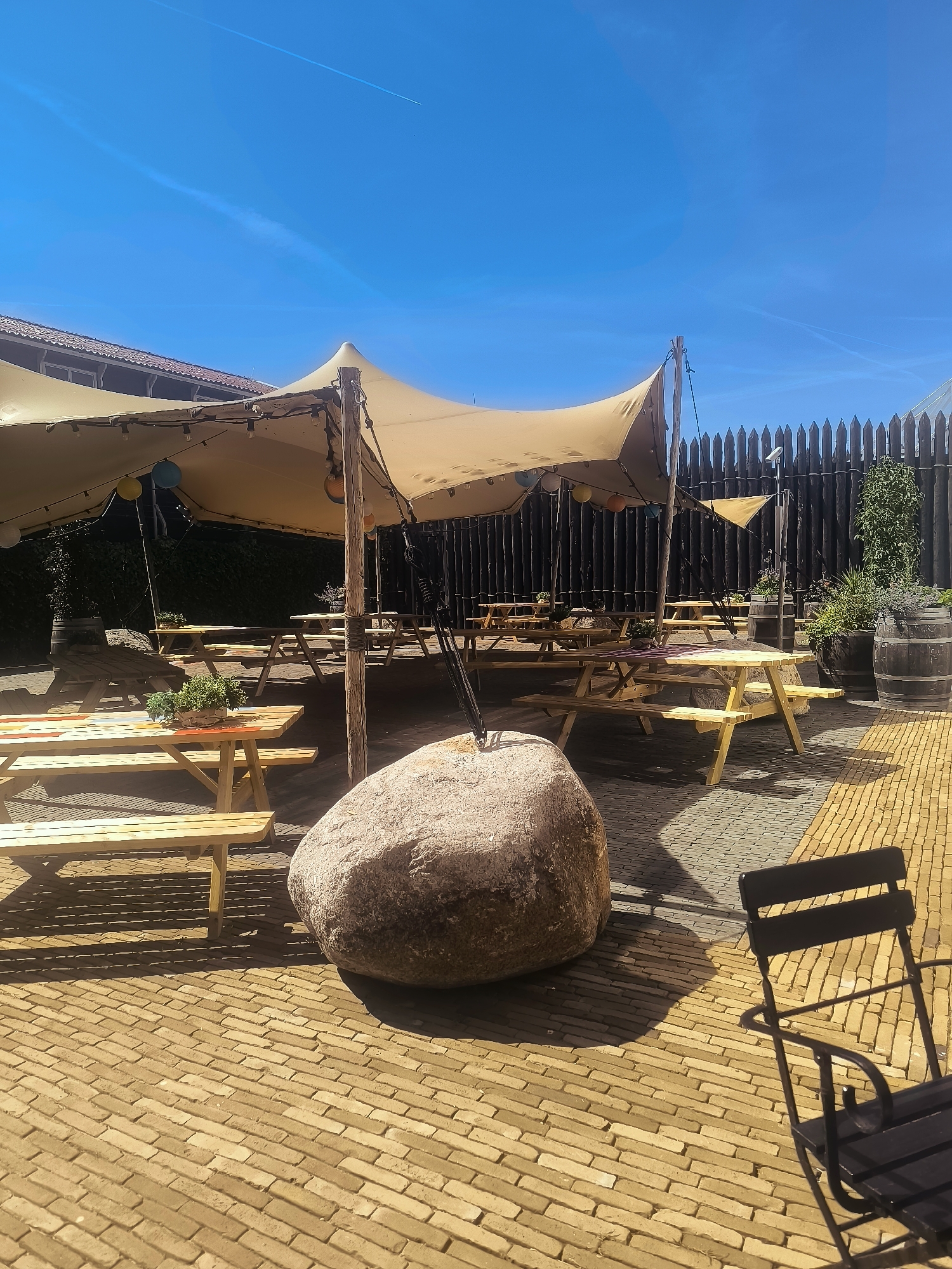 This image captures an outdoor seating area in Lelystad, Netherlands, on a clear, sunny day. The scene is dominated by a large, beige, stretched fabric canopy supported by wooden poles, providing shade over several wooden picnic tables and benches. The tables are arranged on a patio paved with light brown bricks in a herringbone pattern. A large, rough, grey boulder sits prominently in the foreground, casting a shadow on the ground. In the background, a dark wooden fence with a spiked top encloses the area. There are also several large wooden barrels, some containing greenery, and a solitary black metal chair positioned in the lower right corner of the frame. The sky is a vibrant blue with a faint contrail visible. The overall impression is of a relaxed, outdoor dining or gathering space.