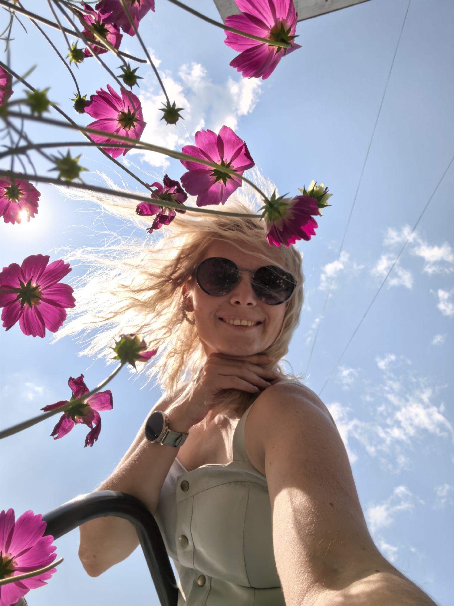 The image, taken from a low angle against a bright blue sky with scattered white clouds, features a woman with blonde hair and sunglasses, smiling. She is framed by several vibrant pink cosmos flowers that appear to be growing overhead. The woman is wearing a light-colored top and a watch on her left wrist. The background suggests an outdoor environment, possibly a park or garden. The location is identified as Sterlitamak, Russia, though no specific city landmarks are visible. The overall impression is one of a sunny day and a cheerful moment captured outdoors.