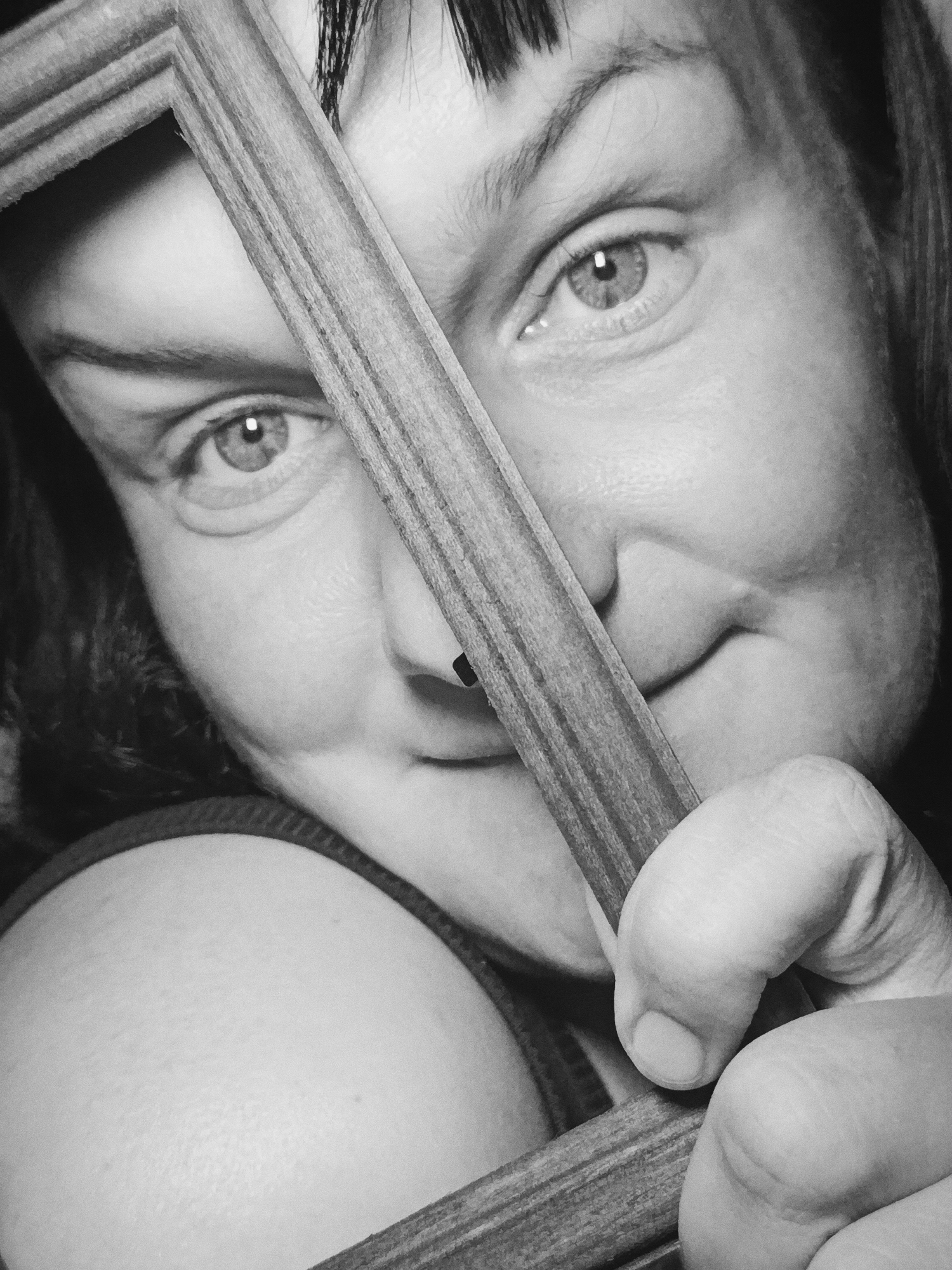 A black and white close-up photograph depicts a person holding a wooden frame partially obscuring their face. The person has short bangs, light-colored eyes, and is smiling slightly. Their right hand, seen holding the frame, shows a closed grip. The background is dark and indistinct. The location and city are unknown.