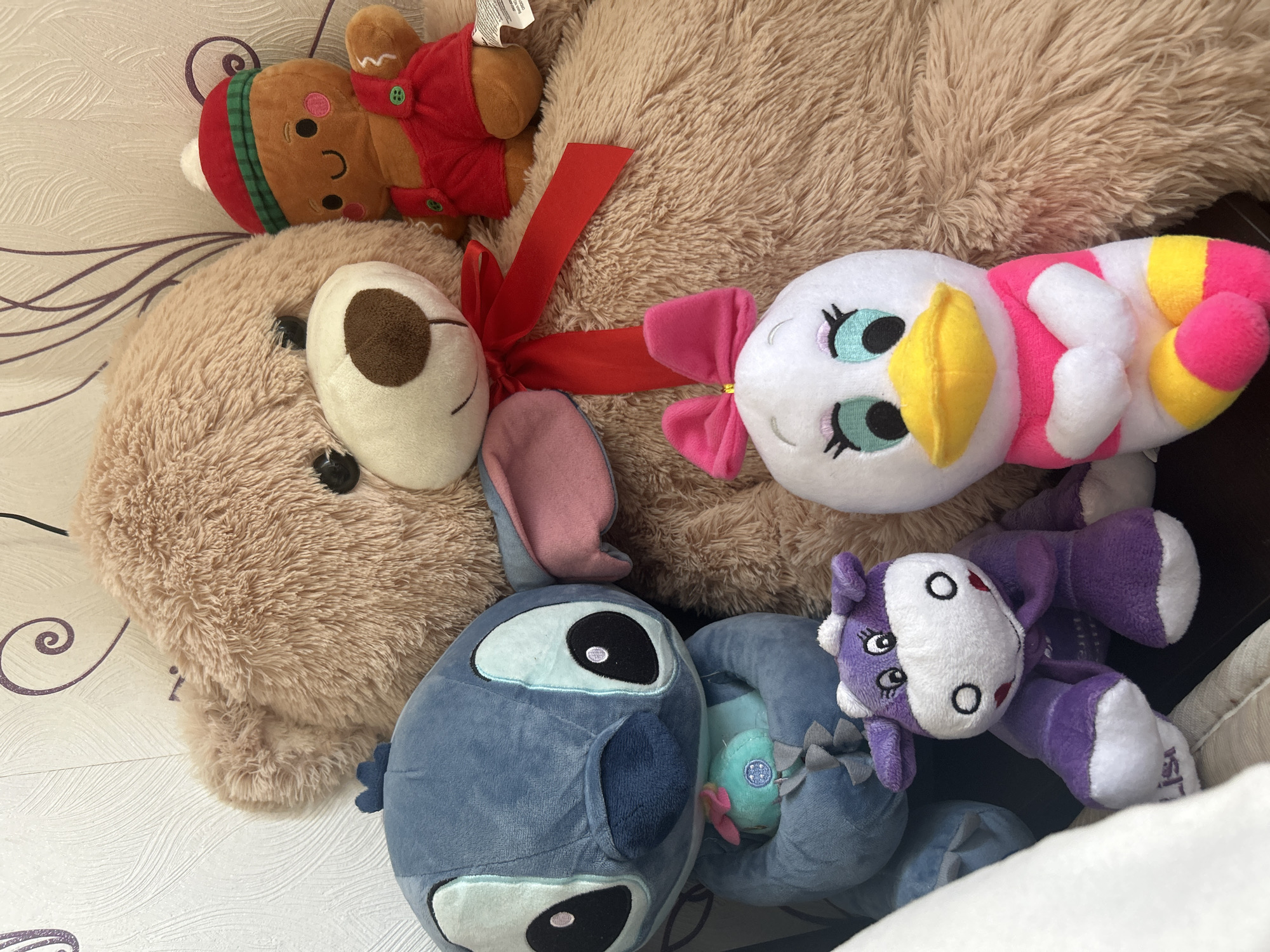 The image shows an overhead shot of several plush toys on a surface with a white patterned background. The toys include a gingerbread man wearing a red vest, a large tan teddy bear with a red ribbon, a white duck with pink and yellow accents, a blue Stitch character with a small light blue plush, and a purple and white horse. The background appears to be a patterned wallpaper or bedding. No people or location are identifiable from the image.