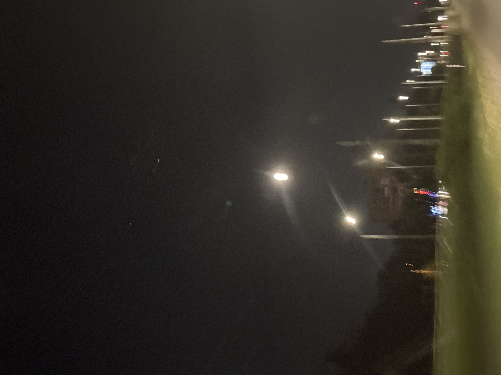 The image shows a nighttime scene taken from a moving vehicle. On the right side, blurred green foliage and a row of streetlights are visible. Further back, blurred city lights, including red and blue lights, can be observed. The left side of the image shows a dark night sky with a few visible stars. The streetlights suggest the presence of a road. The location may be near a park or green area along a road in Sofia, Bulgaria, given the streetlights, city lights, and green space.