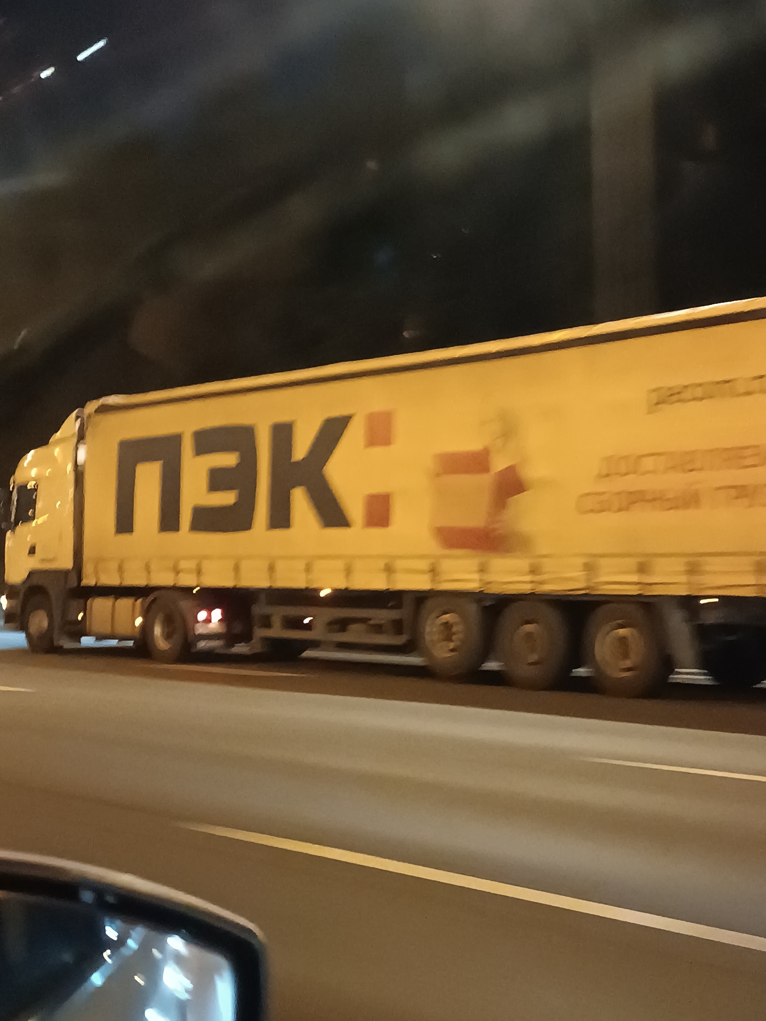 A yellow truck with dark blue lettering "ПЭК" is driving on a highway at night. The truck is large and appears to be a semi-trailer truck. The lettering "ПЭК" is prominently displayed on the side of the trailer. To the right of "ПЭК", there is a graphic of two red boxes stacked on top of each other, with a third red box partially visible to the left. Further to the right, there is text that appears to be in Russian, with the words "ДОСТАВЛЕНИЕ" and "СБОРНЫЙ ГРУЗ" visible. The truck has multiple wheels and appears to be moving at a good speed. The road is dark and appears to be well-lit by streetlights and the truck's headlights. The background is dark and shows a hint of a tunnel or overhead structure. The image is taken from a slightly elevated perspective, possibly from another vehicle. The overall scene suggests a commercial transport operation occurring on a highway.
