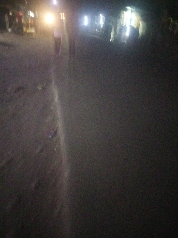 A dimly lit street scene at night. Two figures are walking away from the camera, their forms blurred. Streetlights and vehicle headlights create bright, indistinct pools of light in the background. The road surface is textured and partially obscured by shadows, with possible debris visible on the left side of the road. Overall, the image has a soft focus, contributing to a somewhat dreamy or ethereal atmosphere.