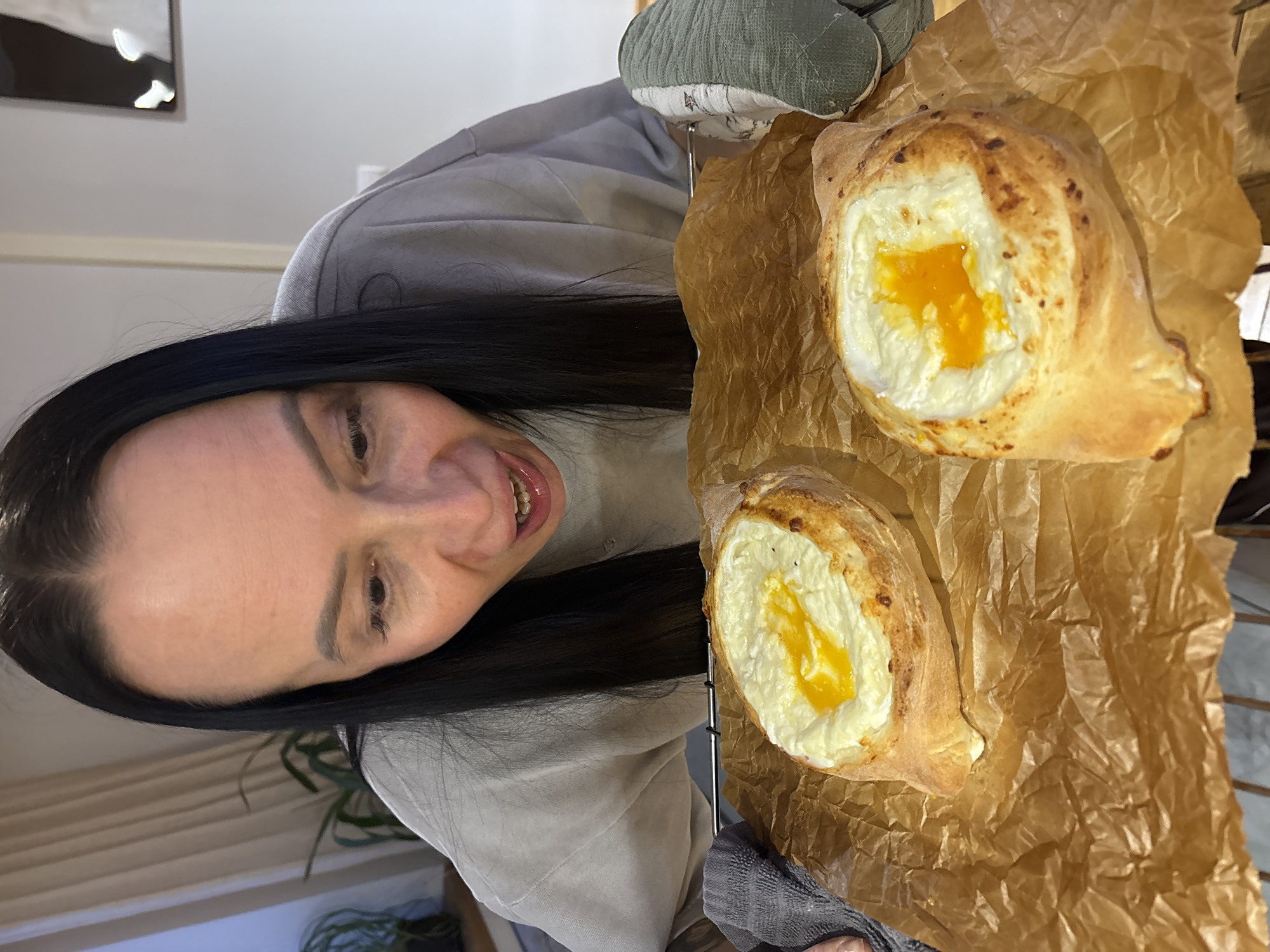 An adult female is depicted indoors in Chaikovsky, Russia, closely observing two freshly baked Khachapuri Adjaruli. The woman, with long dark hair and wearing a grey long-sleeved top, leans forward with an open mouth and an intensely focused gaze directed at the food. She holds a silver metal oven rack, on which two boat-shaped bread items filled with white cheese and a prominent orange-yellow egg yolk are presented atop crumpled brown parchment paper. Her left hand is protected by a grey textured towel, while her right hand is covered by a green and white patterned oven mitt. The background reveals an indoor setting with a window and light-colored curtains to the left, alongside a potted plant, and a plain light-colored wall with a visible dark rectangular object and a light switch/outlet to the right.