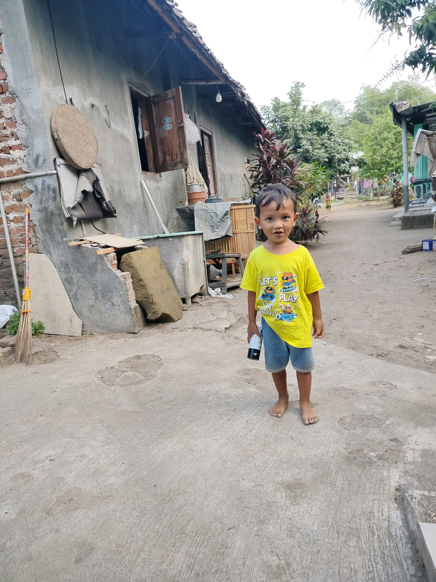 A young boy, appearing to be around four to six years old, stands barefoot on a concrete pathway in front of a modest, weathered building in the village of Beloh, Indonesia. He is wearing a bright yellow t-shirt with a cartoon graphic and blue denim shorts. In his right hand, he holds a dark-colored cylindrical object. The building behind him shows signs of age and repair, with visible brickwork and a corrugated metal roof. A round woven mat hangs on the wall, and various items, including a broom, are scattered around the exterior. Green foliage and trees are visible in the background, suggesting a rural or semi-rural setting. The sky is overcast, and the overall lighting indicates it is daytime.