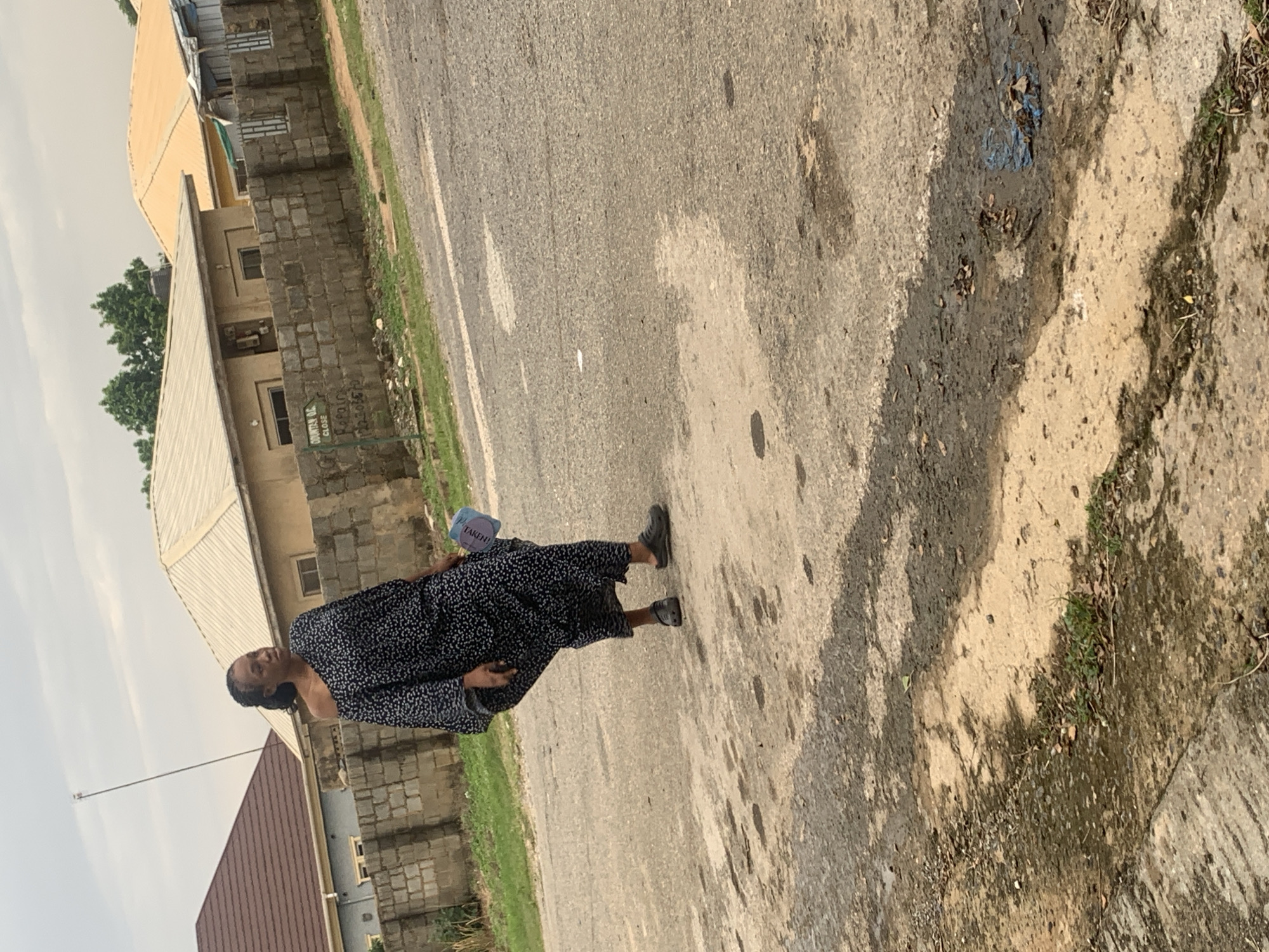 A woman is walking on a paved road in front of a brick wall. She is wearing a dark, off-the-shoulder dress with a white polka dot pattern and dark sandals. She is holding a light blue object in her right hand, which appears to be a small bag or fan. The road is wet in places, with some patches of dirt and debris. Behind the brick wall, there are several houses with different colored roofs, and some trees in the background. The sky is overcast. The location appears to be a residential area in Abuja, Nigeria.
