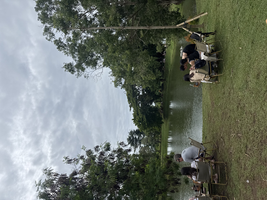 People sit in chairs on green grass beside a pond. Lush trees surround the water, with a cloudy sky overhead. Some people are gathered closer to the water's edge, while others are further back. The chairs are simple folding chairs. The setting has a peaceful, outdoor ambiance.