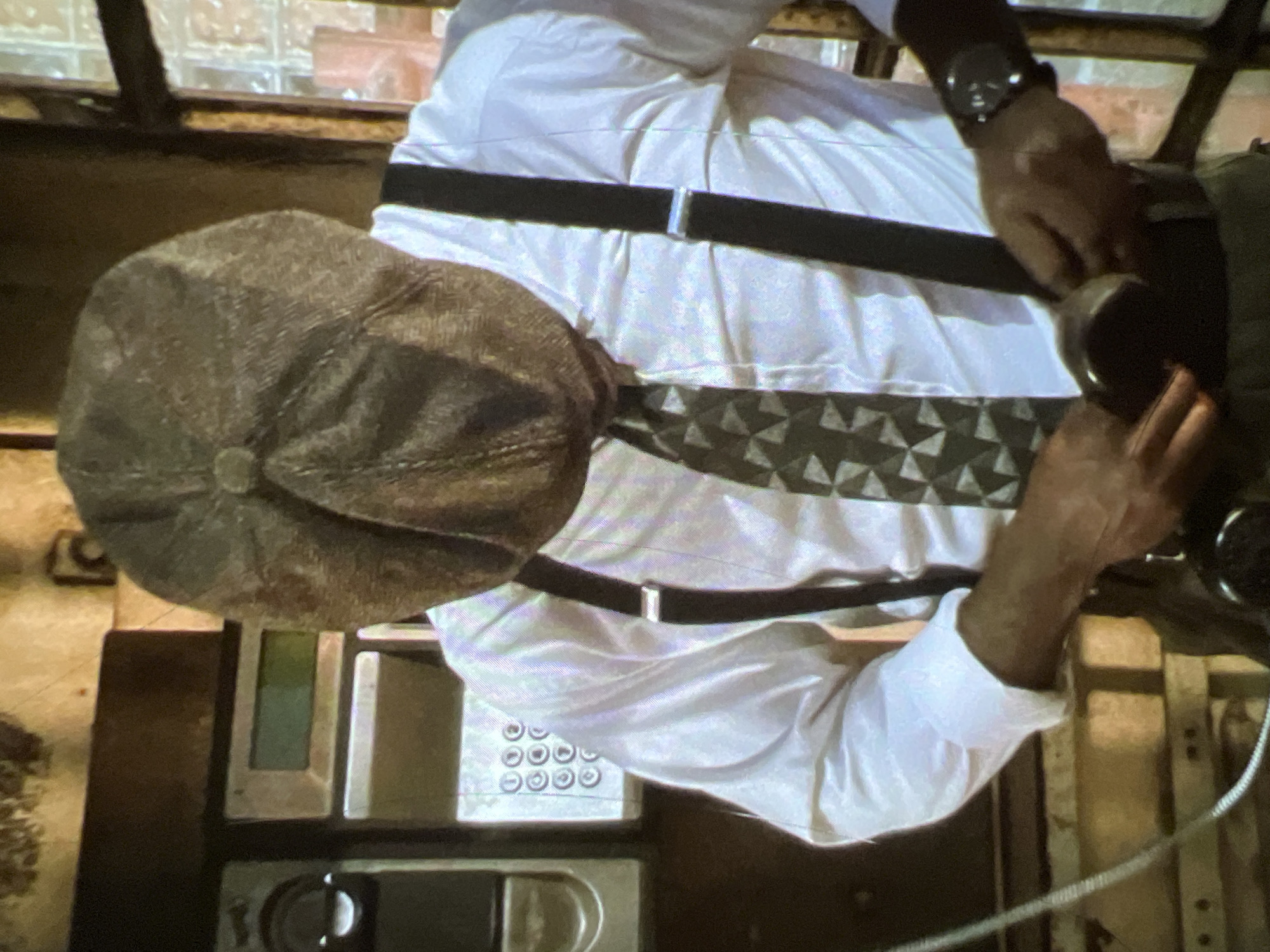 A man wearing a white dress shirt, dark suspenders, a patterned tie, and a flat cap is shown adjusting his suspenders. He is standing in front of a wall with a vintage payphone mounted on it. The payphone has a keypad with circular buttons and a small display screen. The environment appears to be indoors, with a window visible in the background. The man's face is obscured by his cap. The scene does not provide any specific indicators of being in the city of Lagos, Nigeria.