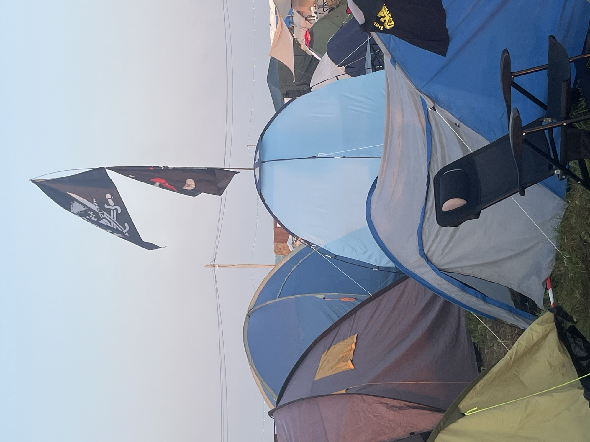 The image depicts a campsite filled with various tents. Two black flags, one featuring a skull and crossbones and another with a red circular emblem, are prominently displayed, flying from a pole above the tents. In the foreground, a black foldable chair is positioned in front of a blue and grey tent. The background shows a cluster of other tents, some with tan awnings. Power lines are visible across the top of the frame, suggesting an outdoor gathering or festival. The sky is a pale, hazy blue, indicating either early morning or late evening light. There are no visible people in the image. The location is identified as Surakovo, Russia.