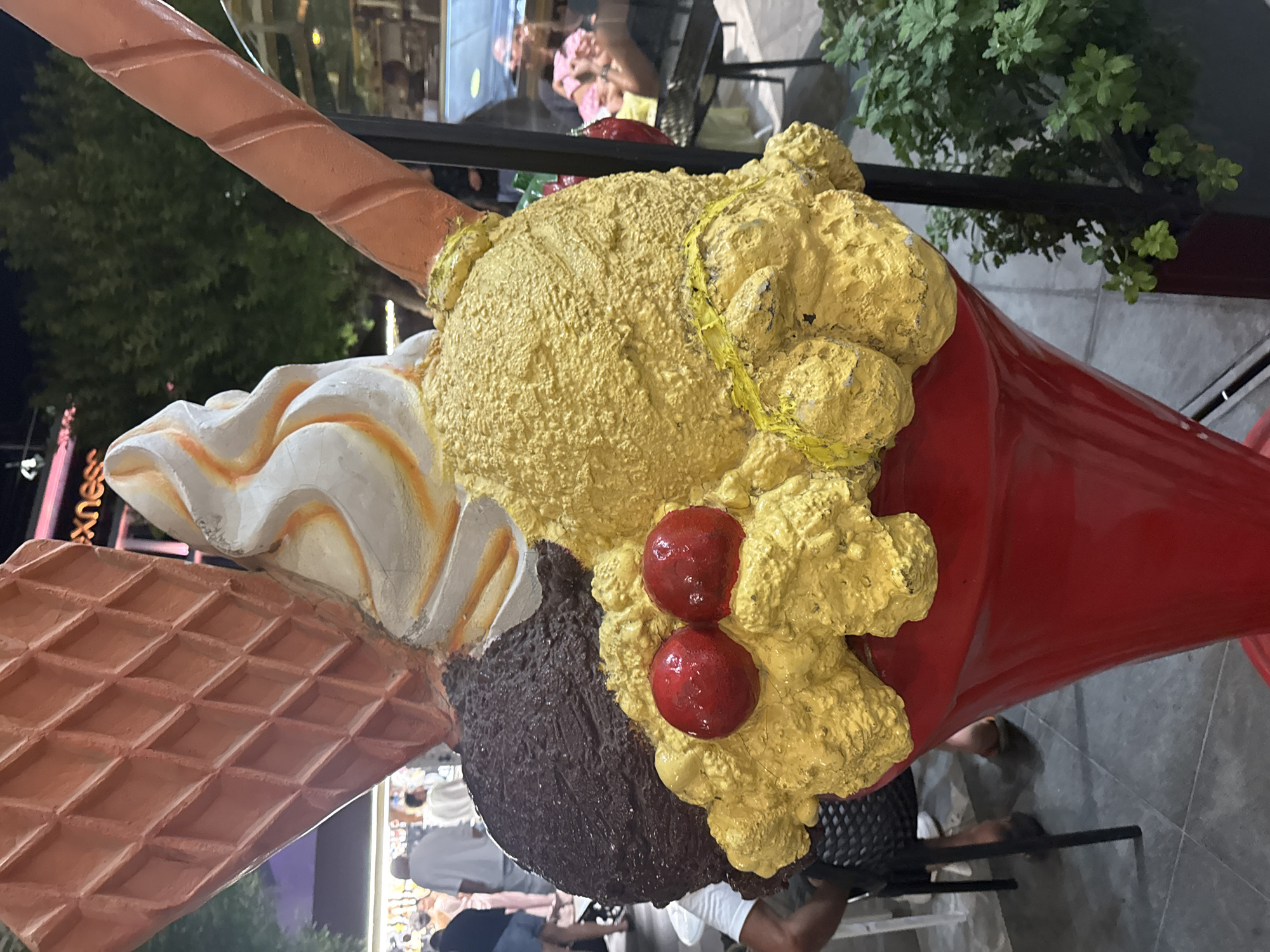 A large, brightly colored, artistic sculpture of an ice cream sundae is prominently displayed outdoors. The sculpture features a red sundae cup overflowing with three large scoops of ice cream: one dark brown, one bright yellow, and one swirled white and orange. Two red cherries sit atop the yellow scoop, and a yellow ribbon is wrapped around it. A large, textured, orange waffle cone section is visible on the left, and a similar, cylindrical orange waffle-textured stick extends from the right side of the sundae. In the background, a blurred urban setting is visible with trees, people walking, and building structures, including a sign with the word "exnes" illuminated. The sculpture appears to be located in a public area, possibly a promenade or a street in Limassol, Cyprus, given the presence of palm trees and the Mediterranean architectural style. The lighting suggests it is either dusk or evening.