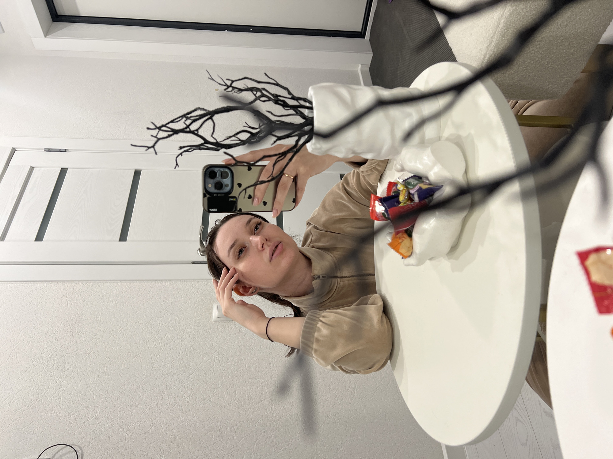 A young woman is taking a mirror selfie in an indoor setting, likely a home in Tyumen, Russia. She is positioned to the left of the frame, looking directly at the camera reflection, with her head slightly tilted and her left hand touching her temple. She wears a casual, light beige or tan zip-up jacket. Her right hand holds a smartphone with a light-colored case adorned with small dark heart shapes; "SNM" is visible on the case, and some inverted text is reflected on the screen.
In the foreground and to the right, a white round table is visible, upon which sits a white decorative vase or container filled with black, gnarled, artificial branches. Another white dish on the table contains an assortment of colorful candies in wrappers. The background reveals a textured white wall, a white door with dark vertical panels, and a light switch. Reflections suggest additional furniture, possibly chairs with textured upholstery, further into the room. The lighting appears bright, indicative of daytime or well-lit interiors. The overall scene suggests a casual moment captured within a modern home environment.