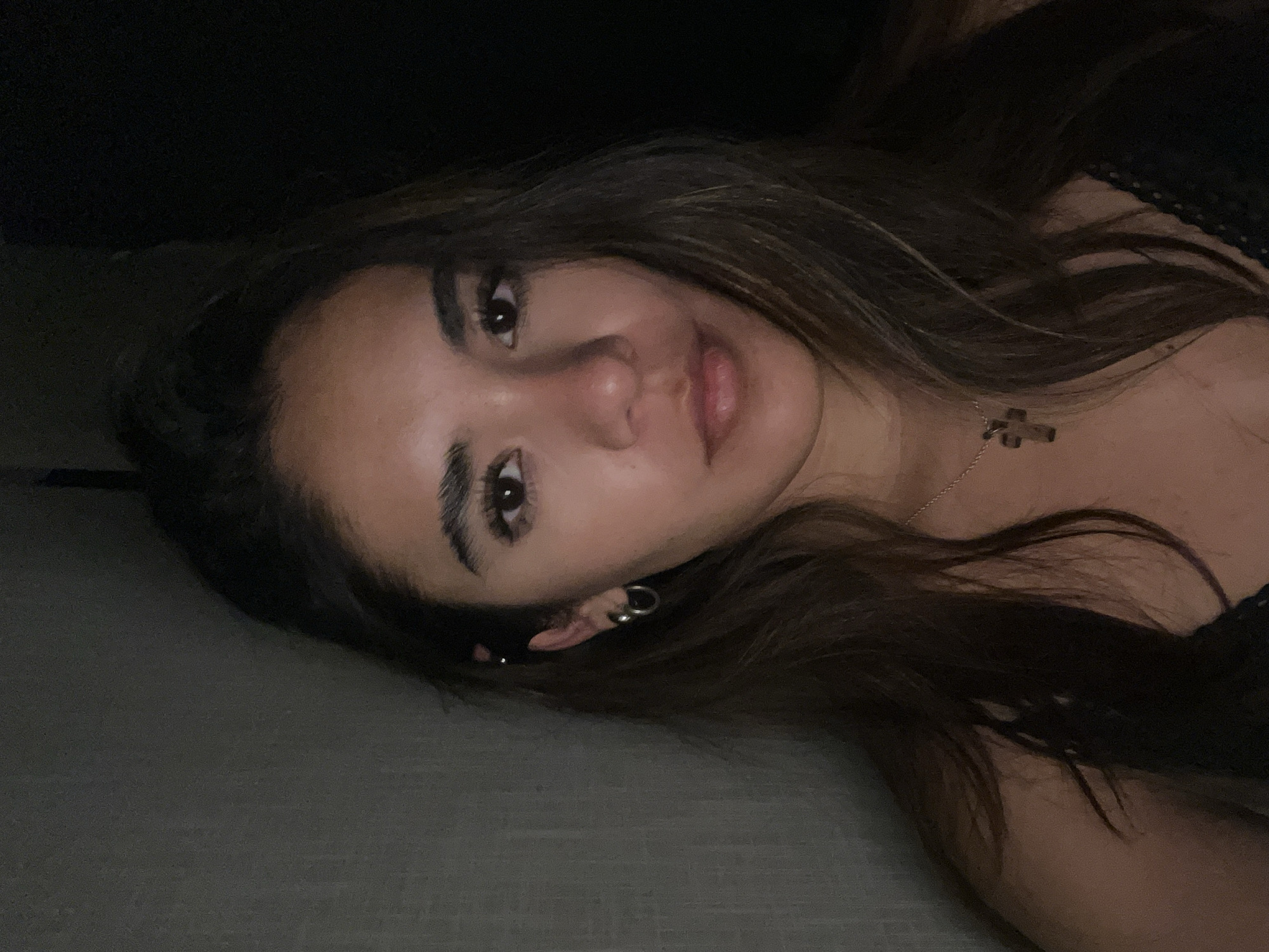 A woman with long brown hair is lying down. She is wearing a necklace with a cross pendant and earrings. The background is dark and undefined, suggesting an indoor setting. The surface she's lying on appears to be a gray or neutral-colored material. There is no visible information to place the image in San Bernardino Tlaxcalancingo, Mexico.