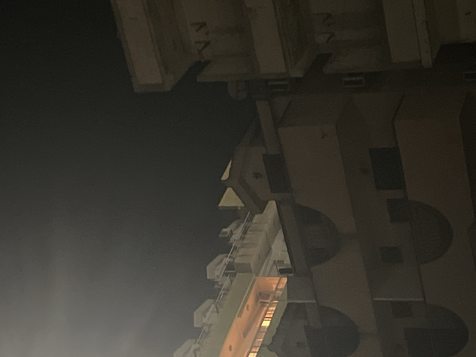 The image captures an upward view of a tall building at night in Budva, Montenegro. The building is predominantly beige with dark, semi-circular balconies. Some windows are visible, with an orange glow emanating from within. The sky is dark and overcast.