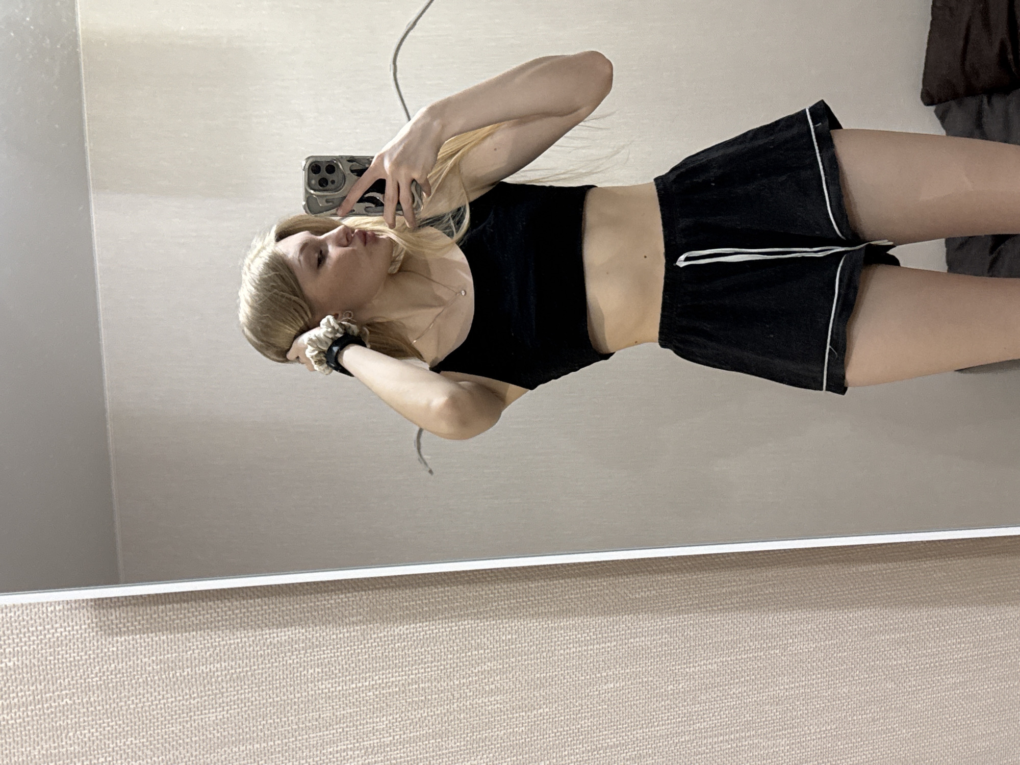 A young woman with blonde hair is taking a selfie in a mirror. She is wearing a black crop top and black shorts with white trim. She is holding her phone in her left hand and has her right hand in her hair. The background is a beige wall. The image may have been taken in Perm, Russia, although the location cannot be confirmed based on the image alone.