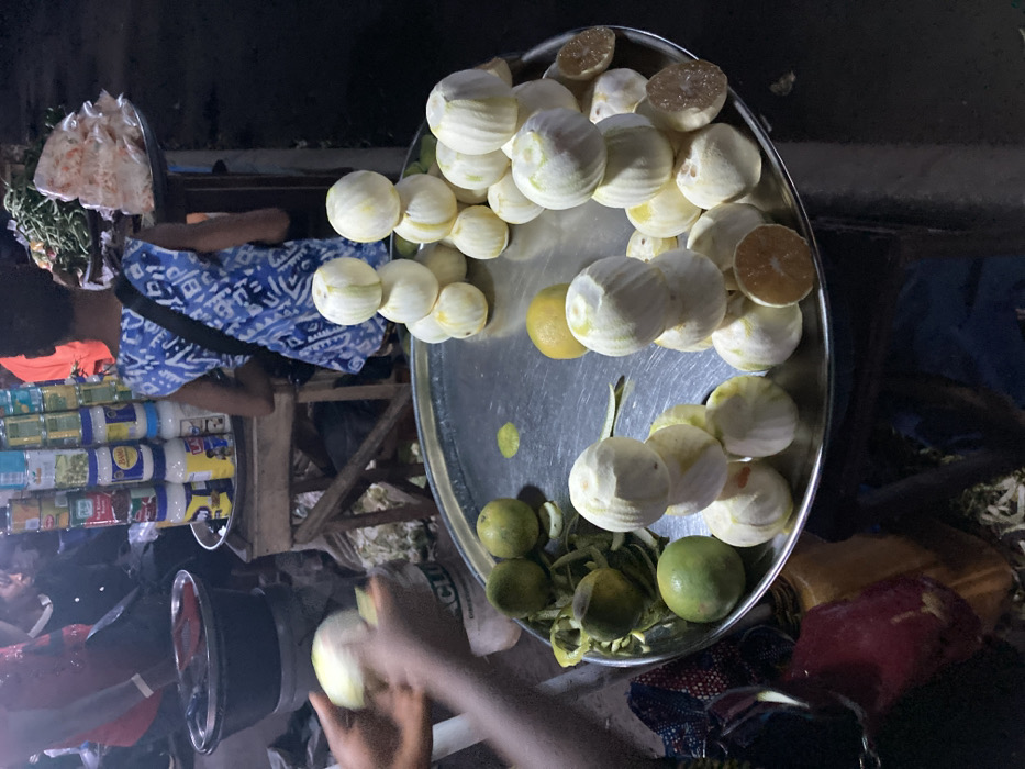 The scene depicts a market activity in Ura Nta Umuarandu, Nigeria, under low light conditions, likely evening or night. In the immediate foreground, a pair of dark-skinned hands is actively peeling a light-colored, spherical fruit over a large, round, metallic tray. The tray contains numerous other fruits, including multiple light-colored, peeled spherical fruits stacked in small pyramids (three visible stacks of approximately three to four fruits each), individual peeled fruits, several unpeeled green citrus fruits, and scattered discarded peels. Two peeled fruits on the tray are cut in half, revealing a pale orange interior.
In the midground, a person, possibly a child or young adult, stands with their back mostly to the camera, wearing a blue and white patterned sleeveless top with a dark strap across the shoulder. This individual faces a stall displaying various consumer goods, including stacks of canned vegetables (e.g., peas) and jars of mayonnaise on a wooden stand. To the right of this stall, another display holds green leafy vegetables and items packaged in clear plastic bags on a tray.
In the left background, a person in a red garment is partially visible, facing right. Above this individual, another person's head and shoulders are dimly seen. A black basin or pot rests on a surface to the left of the canned goods display. The ground surface appears unpaved, with debris visible.
