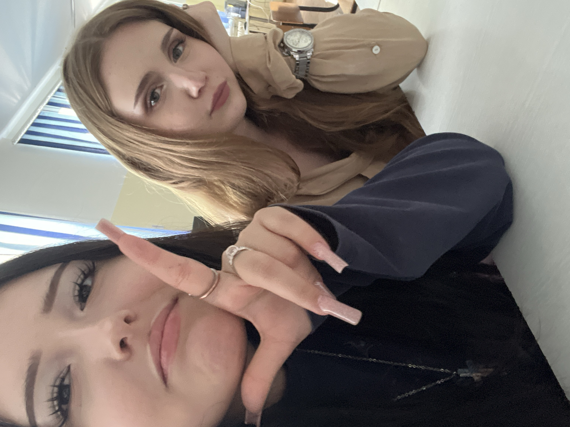 Two young women are seated at a white table, likely in a classroom or common area. The woman on the left is closer to the camera, her face framed by dark hair. She is looking directly at the camera with a slightly pouty expression, her right hand raised, fingers extended with long, light pink acrylic nails. She wears a dark top and a silver necklace with a cross pendant. On her fingers, she has two rings, one plain gold band and a more elaborate silver ring with stones. The woman on the right has long, light brown hair and is looking towards the left. She rests her head on her left hand, which is propped up by her elbow. She is wearing a light brown long-sleeved shirt with a button near the cuff, and a silver wristwatch with a circular face. The background shows a window with blue and white striped blinds, and the corner of a desk or computer monitor is visible on the far right. The scene is well-lit, suggesting it is daytime. No specific landmarks in Sterlitamak, Russia are identifiable from the image.