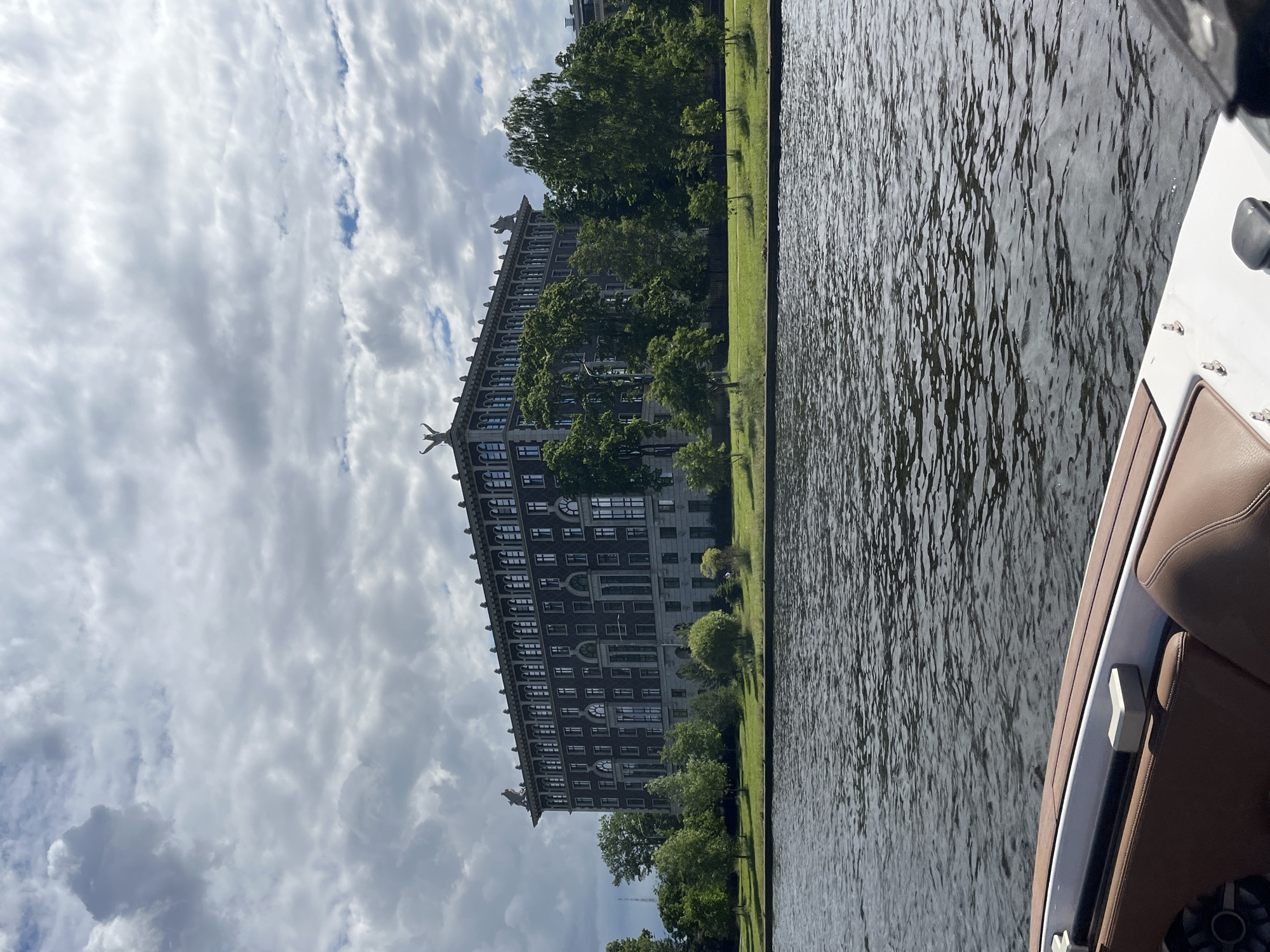 The image depicts a view of the Petrograd Side district in Saint Petersburg, Russia, as seen from the Neva River. In the foreground, the edge of a white and brown boat is visible, with a brown upholstered seat. The river water is dark and rippled, reflecting the overcast sky.
Dominating the background is a large, dark gray, multi-story building with a long facade. It features numerous evenly spaced windows and decorative architectural elements, including statues on its roofline. The building is surrounded by lush green trees and a manicured lawn. The sky above is filled with dramatic, cloudy formations, suggesting an approaching storm or a break in the clouds. The overall scene is one of urban landscape meeting natural elements, viewed from a watercraft.