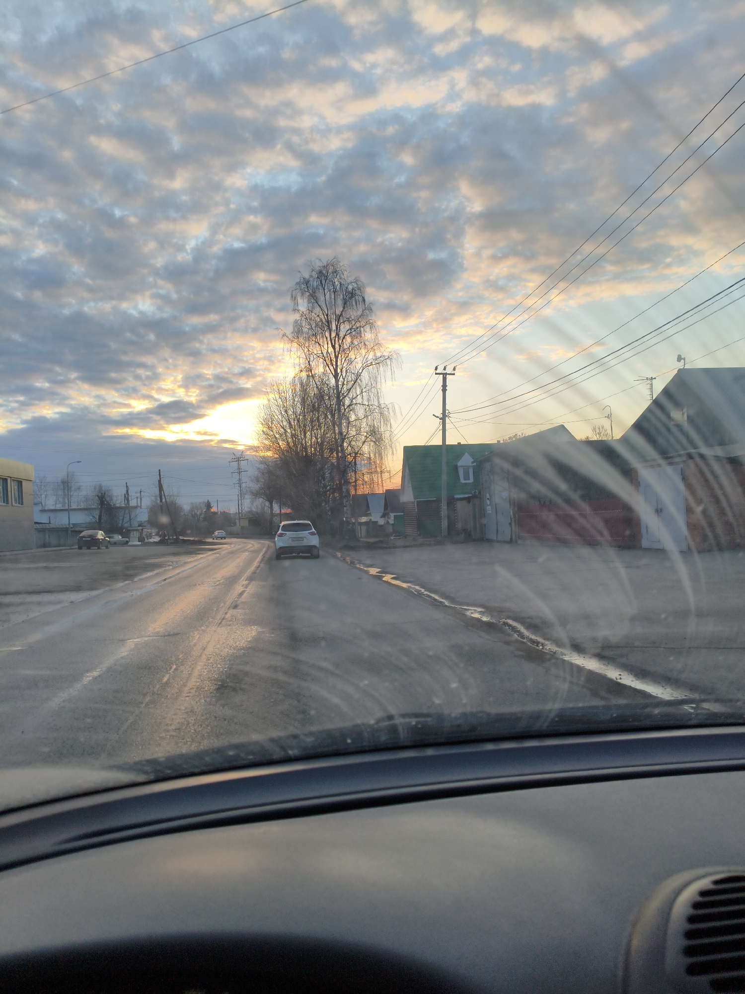 The image, taken from inside a vehicle, shows a street scene in what appears to be the city of Chaikovsky, Russia, during either sunrise or sunset.
The foreground features the dashboard of the car, partially obscuring the view. Looking through the windshield, a wet road stretches into the distance. In the middle ground, a white car is driving away. To the right of the road, there are some buildings, including one with a green upper level and white window frame. Utility poles and wires run overhead. A leafless tree is visible to the left of the car.
The background showcases a cloudy sky with the sun illuminating the horizon. Other buildings and structures are faintly visible in the distance. The overall scene suggests a quiet, possibly residential area.