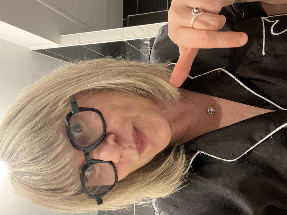 An upright, close-up indoor selfie captures a middle-aged woman with shoulder-length, straight blonde hair and a neutral expression. She is wearing large, black-rimmed eyeglasses, through which her light-colored eyes are visible. Her lips have a soft pink tint.
The woman is dressed in a black satin or silk-like shirt, possibly pajamas, featuring white piping along the collar and front opening. A delicate silver chain necklace with a small, circular pendant adorned with a central embellishment and surrounding details rests on her chest. Her right hand is raised, with the index finger pointing downwards towards her shoulder, seemingly indicating the length of her hair. A silver ring with a dark, tear-drop shaped stone is worn on her right index finger.
The background consists of a modern indoor setting, likely a bathroom or kitchen, featuring black wall tiles with white grout lines, topped by a white border and an upper white wall or ceiling. The lighting is bright and artificial. There is no visible text. The image likely depicts a moment in Chambœuf, France.