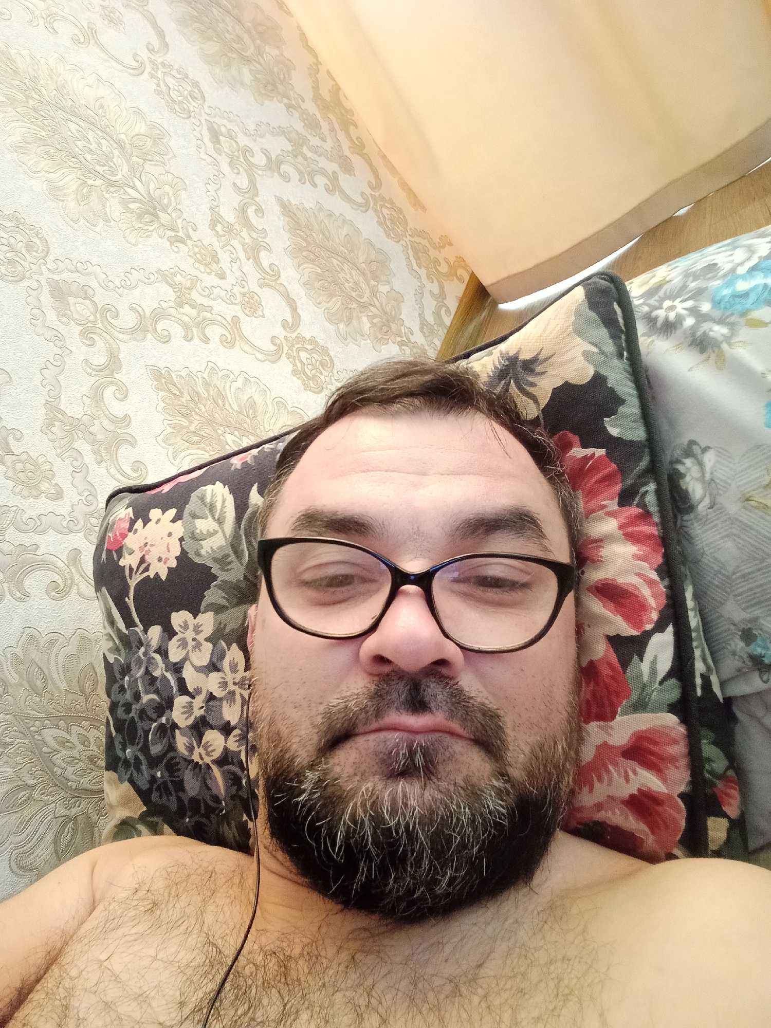 The image is a selfie taken from a slightly high angle, looking down at the subject. The subject, a man with a beard and glasses, is lying down and is shirtless. He is positioned in the foreground, with a floral patterned pillow behind his head. The background consists of patterned wallpaper and a light-colored curtain. The scene appears to be indoors, likely in a bedroom or living area. There is no indication of the location being Samarkand City, Uzbekistan.