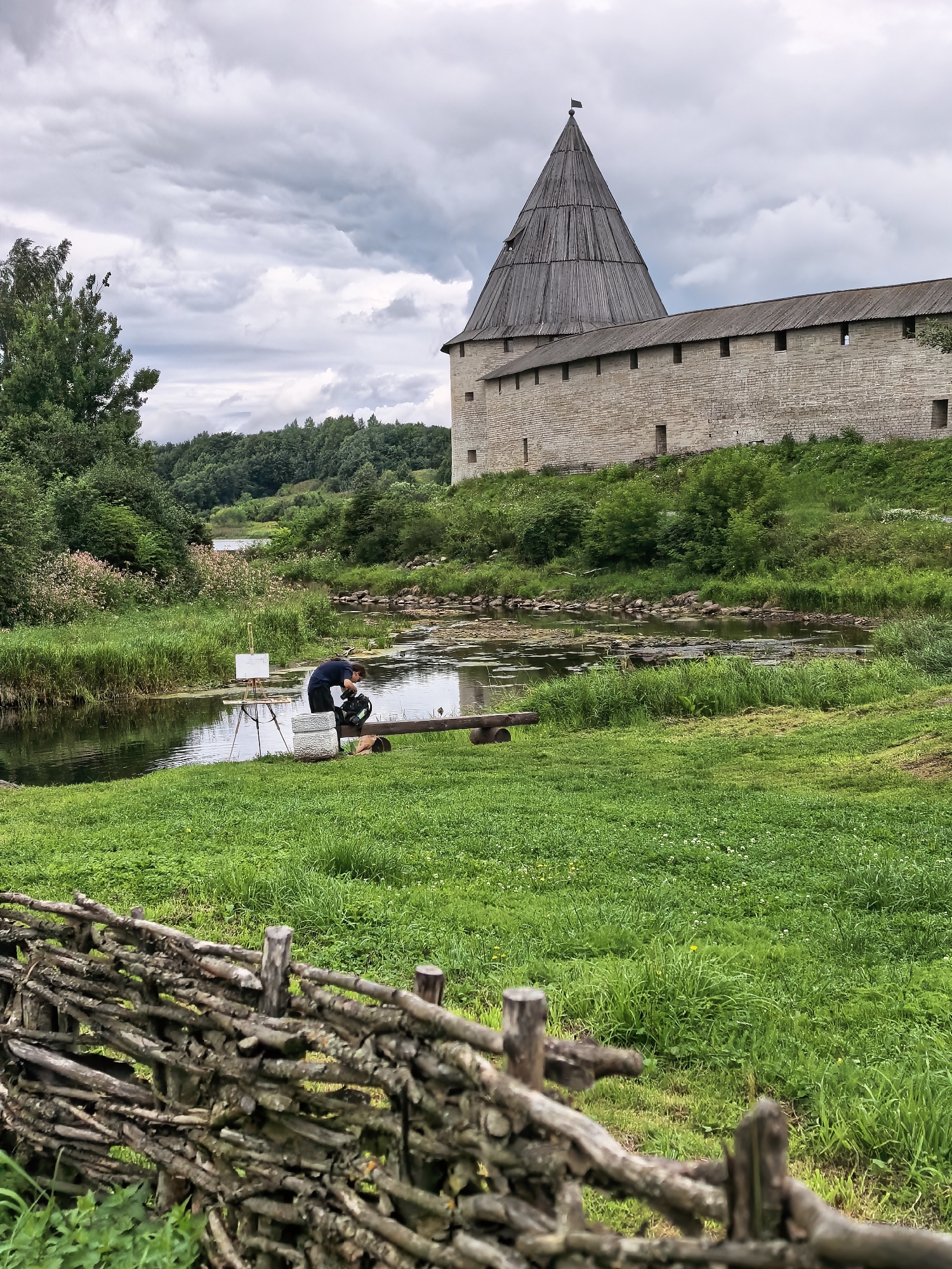 In Staraya Ladoga, Russia, a person is standing by a body of water, seemingly setting up or adjusting a camera on a tripod. The person is wearing dark clothing and is crouched down. Next to them is an easel with a white canvas, suggesting they might be an artist or videographer. The environment is lush and green, with tall grass bordering the water. In the background, a historic stone fortress with a conical wooden roof stands under a cloudy sky. A rustic wooden fence made of branches is in the foreground, adding to the historical and natural ambiance of the scene. The water appears calm, with some lily pads visible. The overall impression is one of an artist capturing the scenic beauty of a historical site.