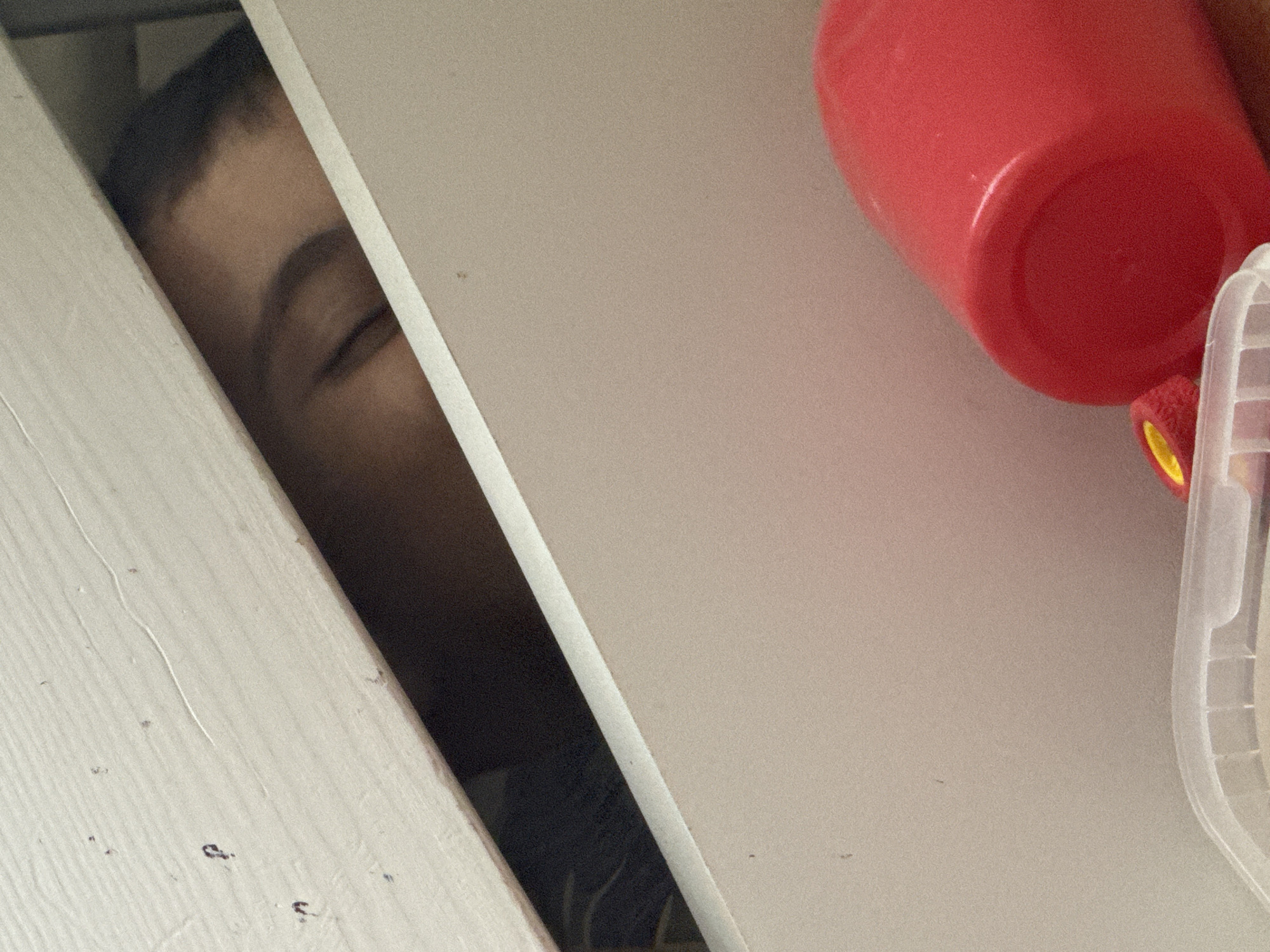 A close-up, high-angle shot shows a partially obscured face of a child peeking through a narrow gap. The child's eye is closed, and the visible skin appears to be light brown. The gap is formed by two white wooden surfaces. To the right of the gap, there's a blurred red plastic cup and a partially visible clear plastic container with a yellow object nearby. The wooden surface on the left has a slightly aged appearance with minor imperfections and texture. The background is mostly obscured. No information about the location in the city of Chaikovsky, Russia is available in the picture.