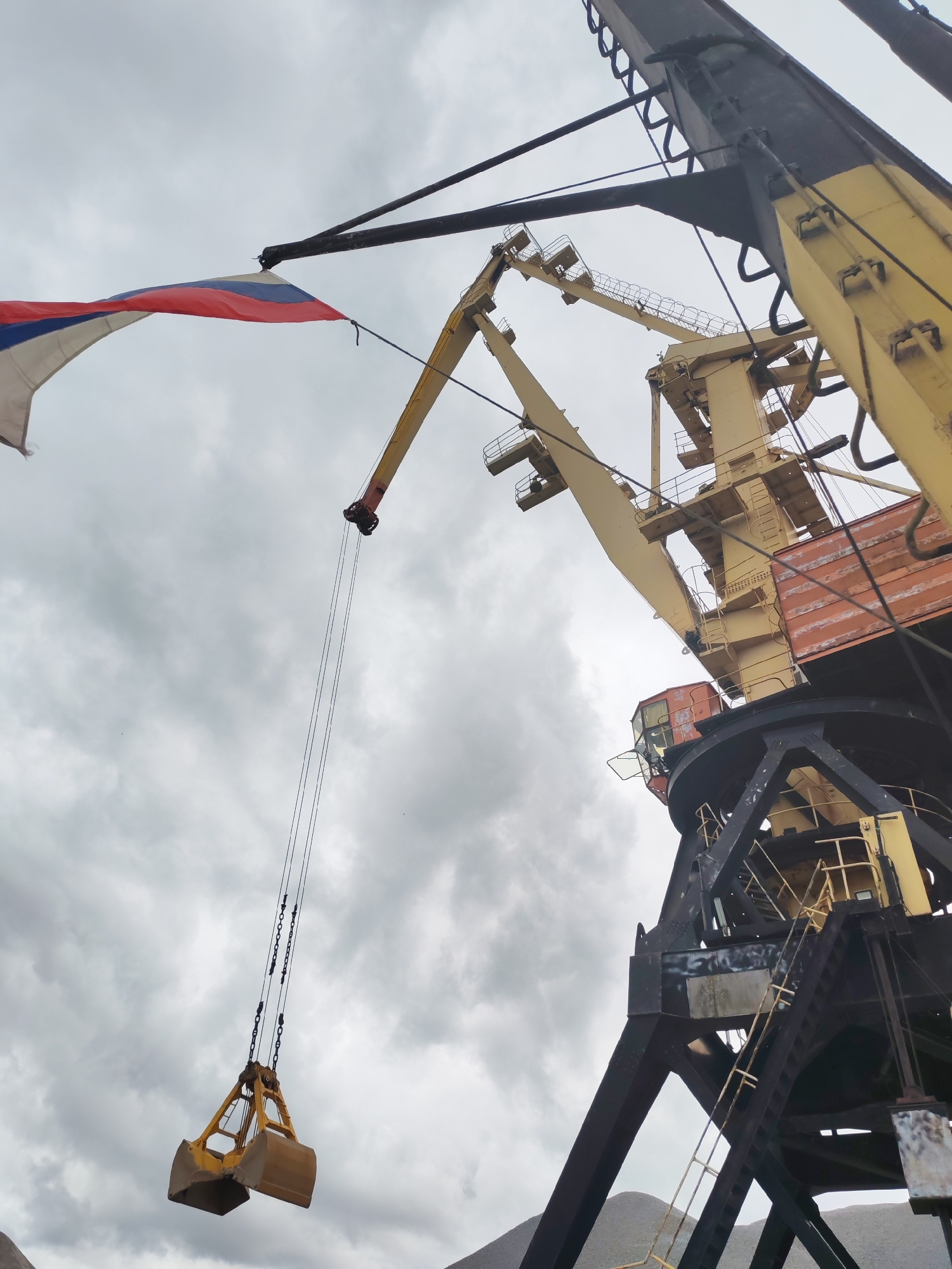 In Bol'shie Mkhi, Russia, a large yellow and black industrial crane stands against a cloudy sky. The crane has a yellow boom extending upwards, and a grab bucket is suspended from cables. A Russian flag is visible attached to a wire on the crane, fluttering in the wind. The base of the crane is a complex structure of black metal beams. In the background, piles of grey material, possibly gravel or stone, are visible, and distant mountains can be seen under the overcast sky. The scene appears to be a port or industrial site.
