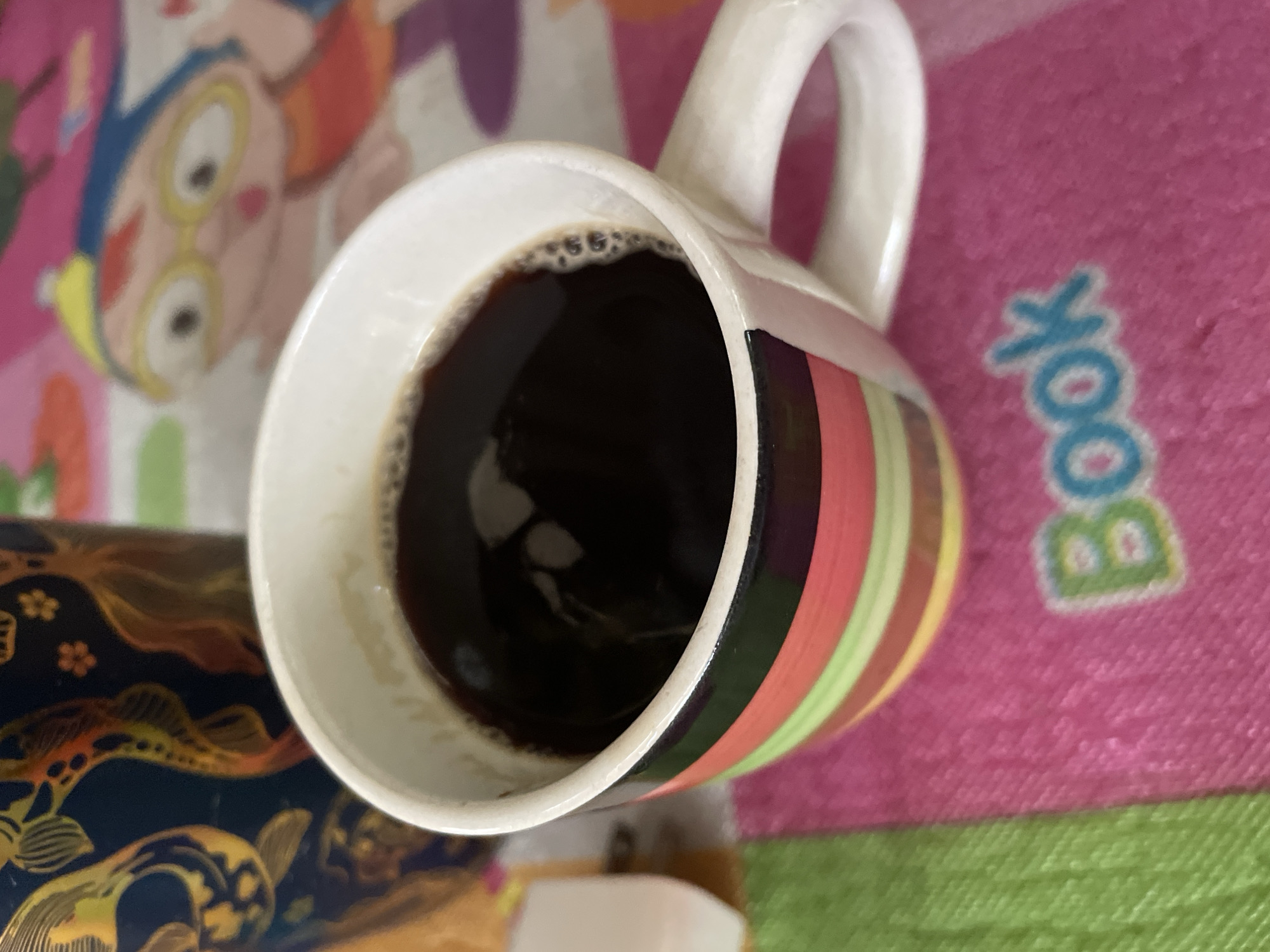 A high-angle shot depicts a colorful coffee mug filled with dark liquid, likely coffee, resting on a multi-colored surface. The mug is mostly white, with a vertical series of stripes in shades of pink, red, green, and yellow. A white handle is attached to the mug. A portion of a decorative object with a dark blue base and gold embellishments is visible at the bottom left. The background surface features cartoon-like figures in bright colors, including pink, green, and yellow. There is a text "book" printed in large blue letters on the pink surface. There is no verifiable information linking this image to the city of Hijo, Philippines.