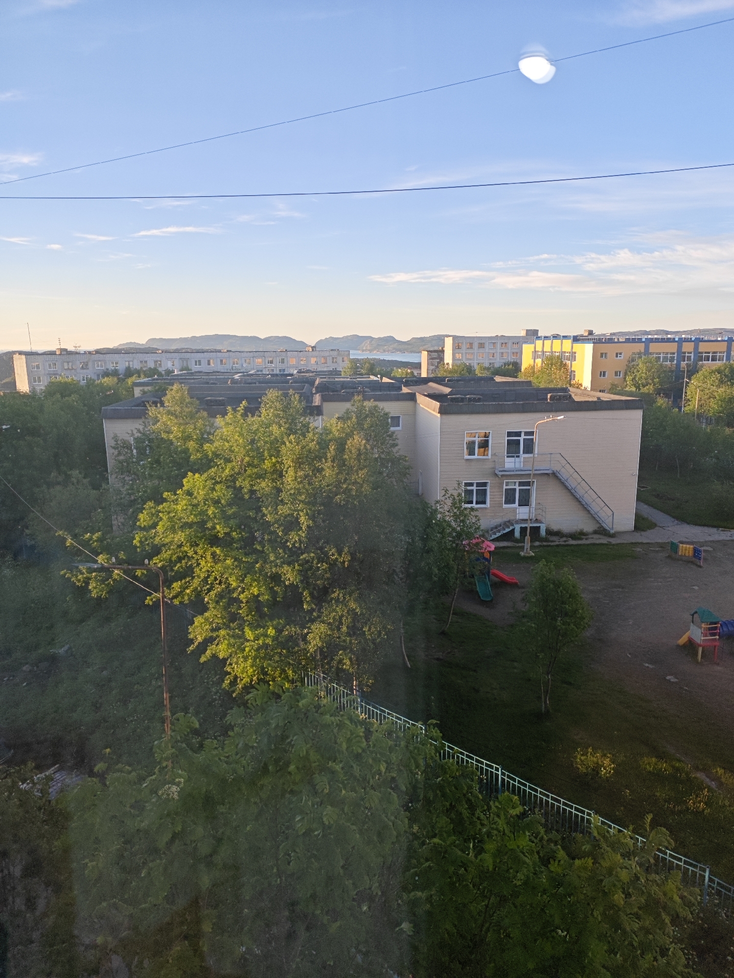 The image shows a residential area in the city of Polyarny, Russia, under a clear blue sky with a few wispy clouds. The sun is low in the sky, casting long shadows and illuminating the scene with a warm, golden light. In the foreground, lush green trees and bushes dominate the landscape, with a white metal fence running along a grassy slope.
A beige, two-story building, likely a kindergarten or community center, stands in the middle ground. It has white-framed windows and an external metal staircase leading to the second floor. Adjacent to the building is a small playground with colorful slides and climbing structures.
In the background, several multi-story apartment buildings stretch across the horizon. Some are lighter in color with visible windows reflecting the sunlight, while others are darker. A body of water and distant, rugged hills are visible beyond the buildings, suggesting the area's proximity to the coast. A single telephone pole with wires is also present in the scene. The overall impression is one of a quiet, sun-drenched day in a northern town.