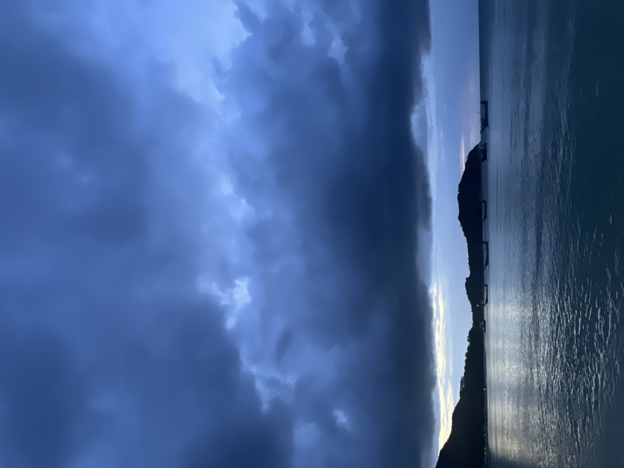 A twilight scene unfolds over the Gulf of Thailand near Ko Pha-ngan District, Thailand, with heavy, dark blue storm clouds dominating the sky. The clouds are dense and textured, with lighter patches suggesting breaks or thinner areas where light attempts to penetrate. Below the ominous sky, the calm sea reflects the dim light, creating a shimmering surface. Several long, narrow boats are scattered across the water, silhouetted against the horizon. On the left and right sides of the image, dark, mountainous landforms rise from the sea, their outlines softened by the fading light. The overall atmosphere is moody and dramatic, indicative of an approaching storm or the aftermath of one.