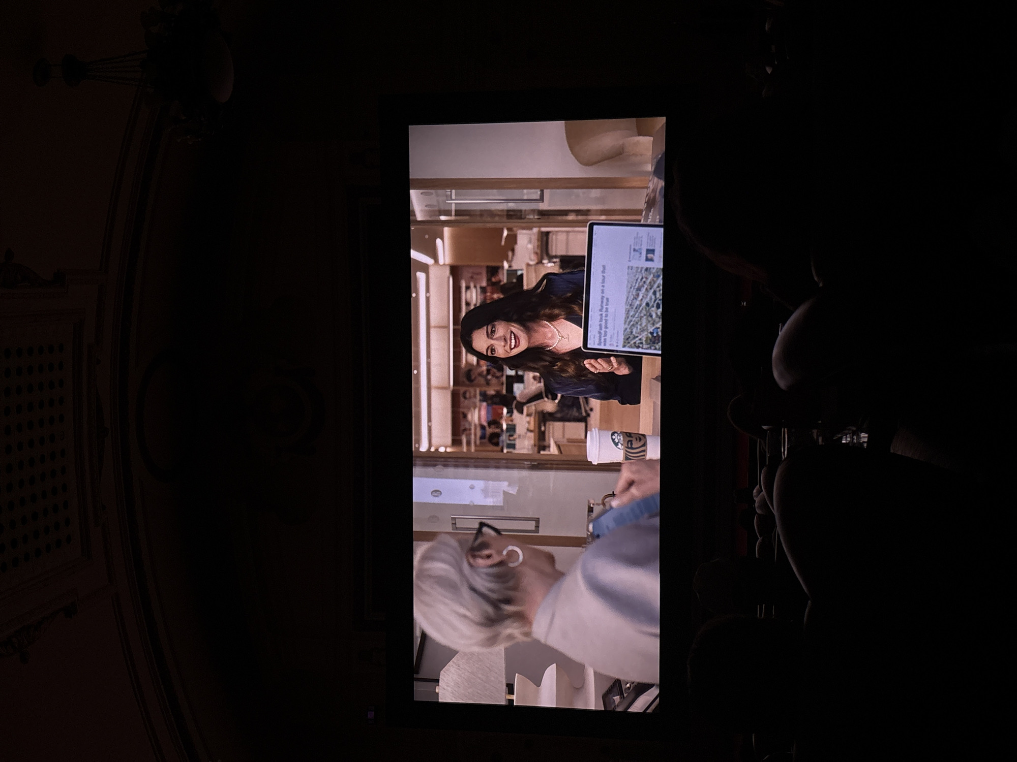 Here is a forensic description of the image:
**Scene:** The image appears to be taken in a darkened room, focusing on a television screen displaying a scene. The screen shows a well-lit interior space that seems to be a high-end retail or department store.
**People:** On the screen, there are two women visible. One woman is holding up a tablet that shows a news headline, and appears to be in conversation with the other woman.
**Objects:** The notable objects include a flat screen television, a tablet displaying a news article, and what appears to be a Starbucks coffee cup. Additionally, there is ornate detailing on the walls surrounding the screen suggesting that it might be a historic theatre or building.