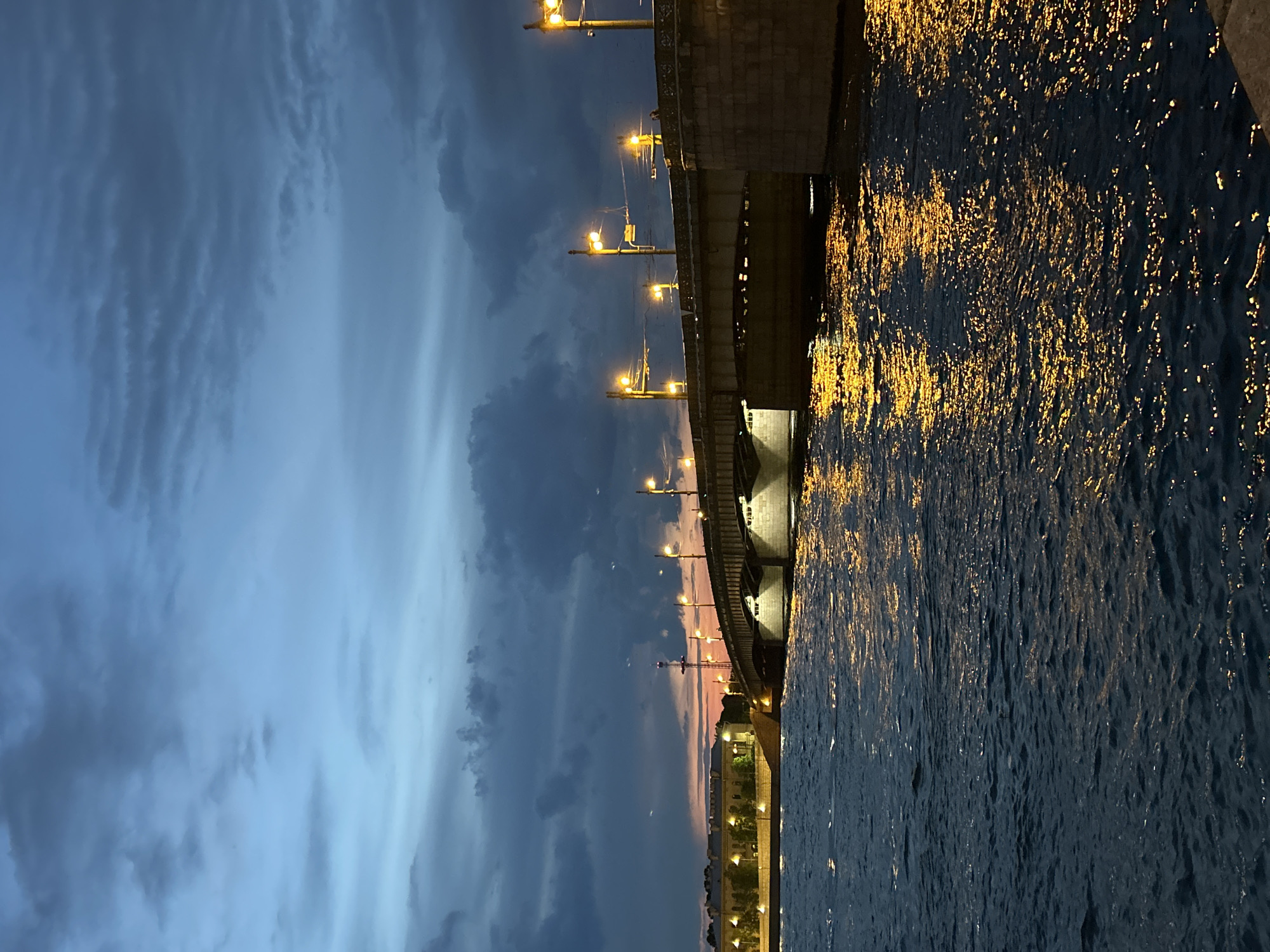 The image displays a dusk or nighttime urban waterscape. A wide body of dark blue water, appearing as a river, occupies the lower portion of the frame, exhibiting ripples and numerous scattered reflections of warm yellow-orange artificial lights.
A multi-span bridge, constructed from stone and metal, extends from the mid-right across the water towards the mid-left. The bridge's deck is lined with a series of tall poles supporting bright, warm-toned streetlights, which cast significant light onto the bridge structure and reflections onto the water below. Additional lighting illuminates the underside of the bridge's arches.
The sky is a dark, deep blue-grey, heavily covered with textured, stratocumulus-like clouds. A narrow band of orange and pink light is visible low on the horizon to the left, indicating the last light of sunset or twilight.
On the left bank of the river, a stone embankment with streetlights lines the shore. Behind this, several multi-story buildings, also illuminated, are visible. A tall, slender, dark structure, resembling an antenna or spire, rises prominently above these buildings against the twilight sky. No individuals are clearly discernible within the scene.
