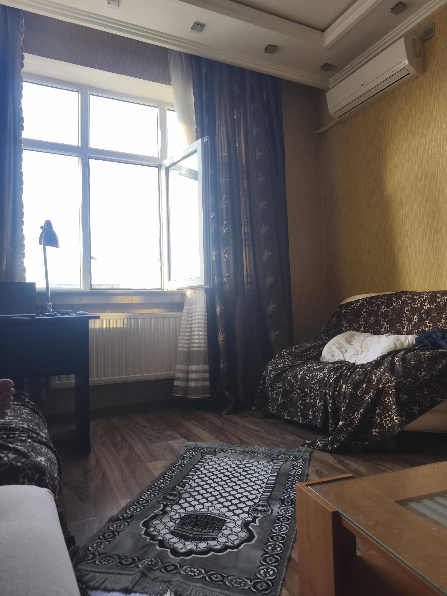 The media file depicts an indoor scene, likely a living room or bedroom in a home in Baku, Azerbaijan. The room is furnished with a sofa covered in a leopard-print throw, a dark wooden desk with a lamp and a laptop, and a wooden coffee table with a glass top. A prayer rug is laid out on the wooden floor in front of the sofa. Large windows with dark patterned curtains and sheer white curtains allow natural light to enter the room, suggesting it is daytime, possibly late morning or afternoon, given the brightness. An air conditioning unit is mounted on the wall near the ceiling. The overall impression is one of a lived-in space, with a sense of comfort and routine, indicated by the prayer rug and the presence of personal items like the laptop. There is no clear indication of any specific activity taking place, but the setting suggests a private, domestic environment. The wallpaper has a textured, yellow-gold pattern.