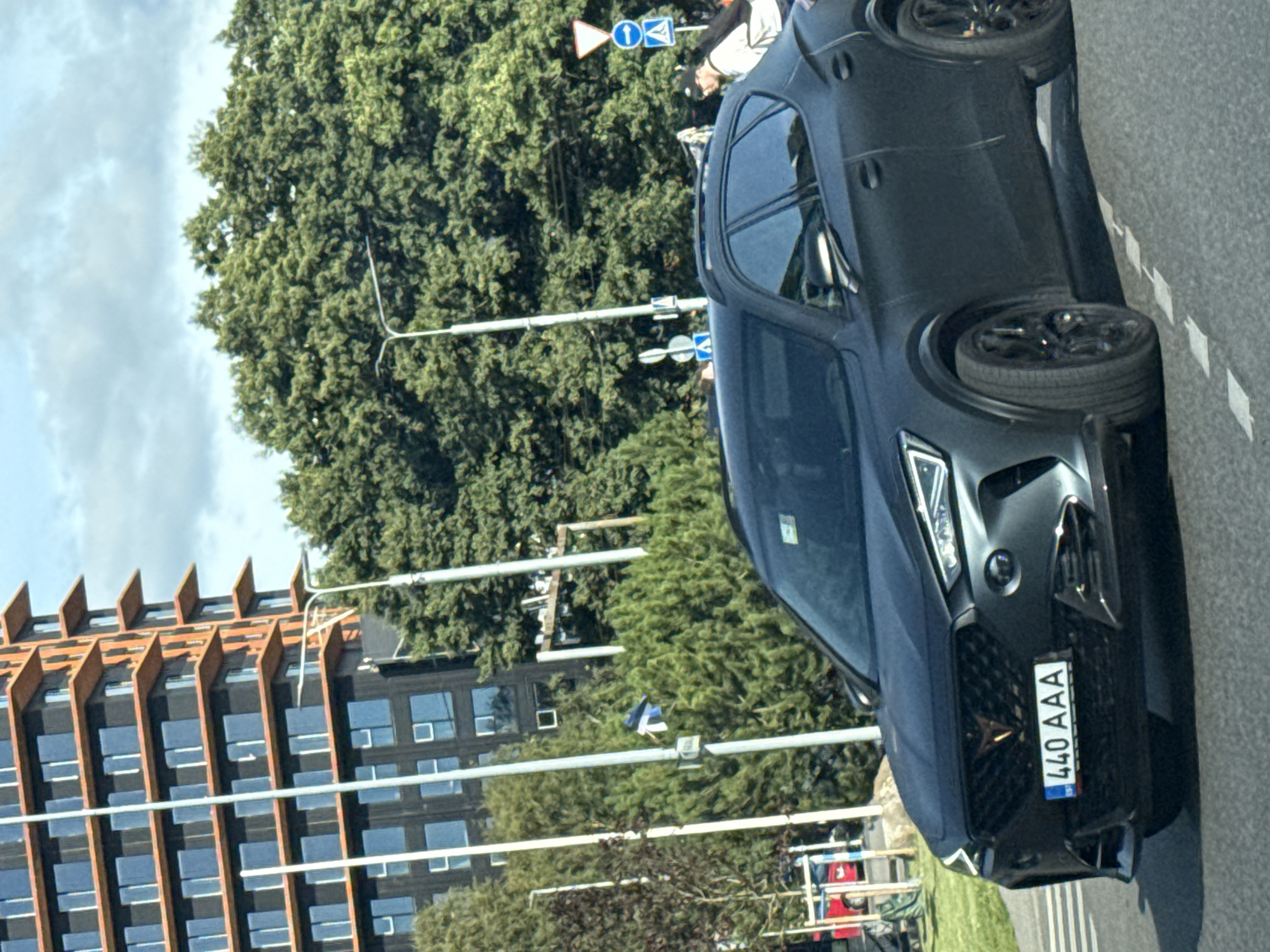 A dark grey coupe is driving on a paved road in Tallinn, Estonia. The car has a license plate that reads "440 AAA". In the background, a modern building with many windows and orange accents stands tall. Lush green trees form a dense backdrop to the scene, with streetlights and traffic signs visible. The sky is partly cloudy.