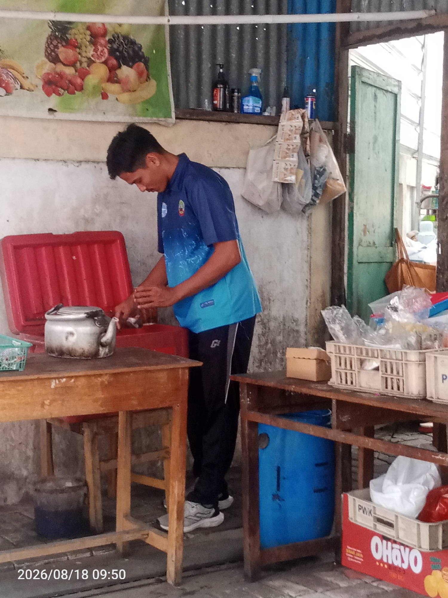 A man in a blue shirt and black pants is standing by a wooden table. On the table is a silver kettle and a red cooler. In front of the table, there is a blue barrel. To the right of the table, there are several plastic crates filled with items. A green wooden door is visible in the background. The scene appears to be an outdoor market stall in Surakarta, Indonesia, suggested by the timestamp "2026/08/18 09:50" and typical market elements like a stall, cooler, and crates of goods. The man is actively engaged in a task, possibly preparing or serving something from the cooler.