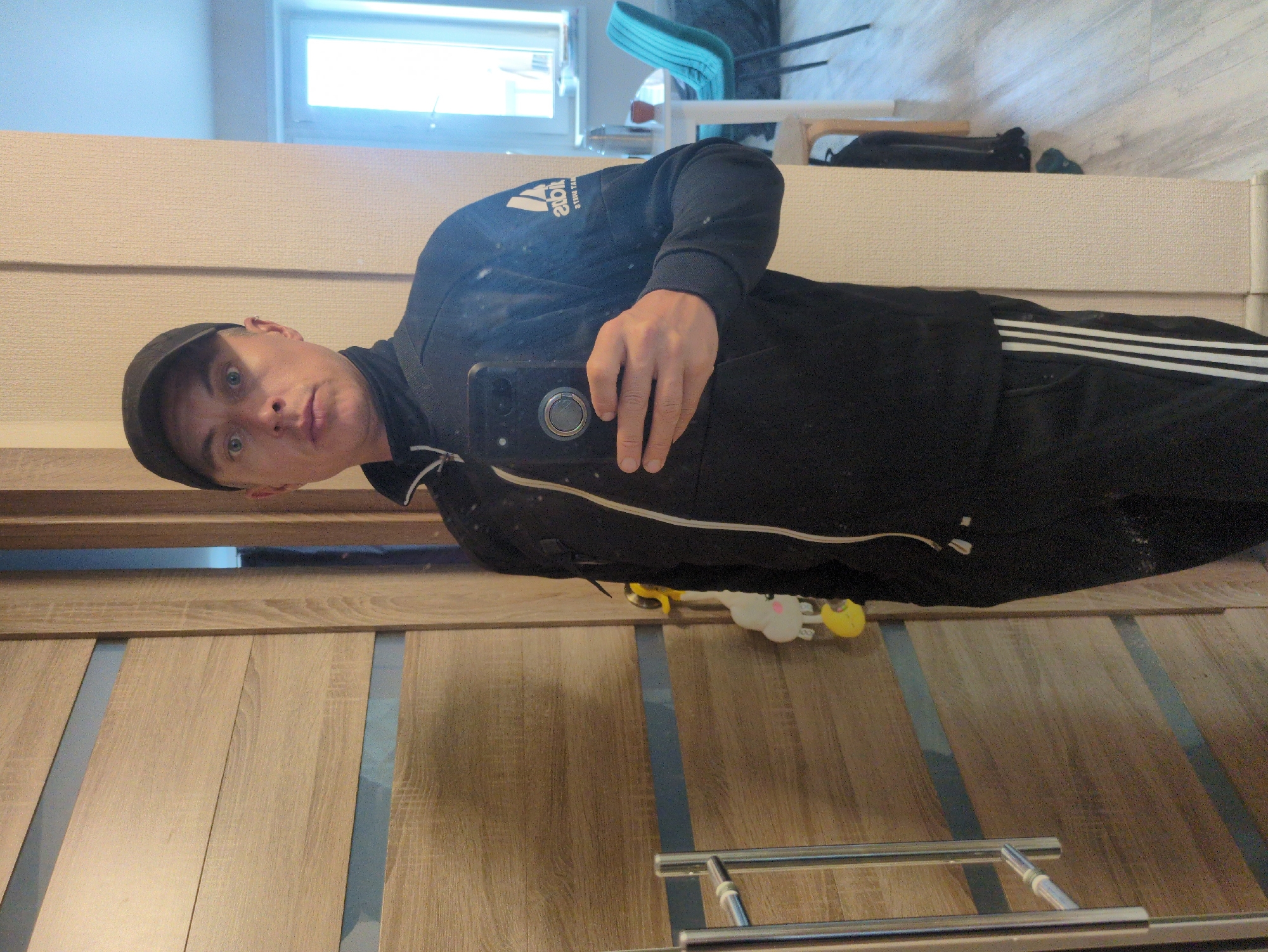 The image is an indoor, eye-level, medium shot of a person taking a selfie in a mirror. The person is wearing a black baseball cap, a black Adidas tracksuit jacket with white stripes on the sleeve, and dark pants. They are holding a smartphone in their right hand, positioned to capture their reflection. The person's face is visible, and they appear to be looking directly at the camera with a neutral expression.
The background shows a well-lit room with what appears to be a large mirror on the wall, reflecting the person and their surroundings. To the left of the mirror, a window is visible, letting in natural light. In the reflection, a white textured wall can be seen above what looks like a cabinet or wardrobe. The flooring is made of light-colored wood planks with darker vertical strips.
On the floor in front of the cabinet, a few small, colorful toys are visible, including a yellow duck and a white figure with pink ears. The overall scene suggests a casual, domestic environment, possibly a home or apartment. The lighting is bright, indicating it is daytime. No specific time of day or weather conditions are discernible. The image contains text on the person's jacket, which appears to be the Adidas logo with text in a mirrored orientation.