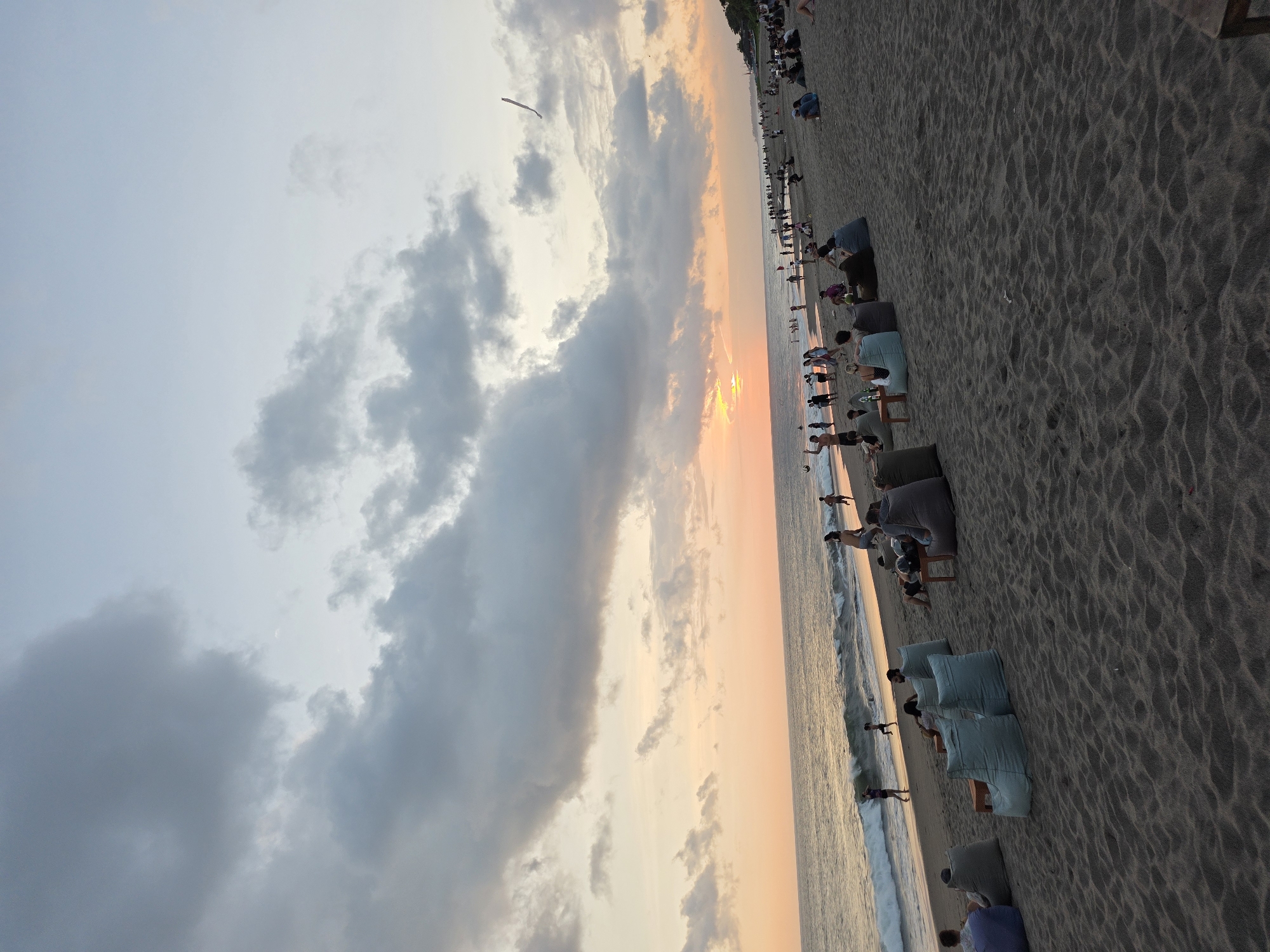 The image depicts a beach scene at sunset, likely in North Kuta, Indonesia. The beach is crowded with people; some are walking along the shore, others are seated in lounge chairs positioned near the water's edge. The sky is filled with clouds and colored by the setting sun, creating an orange and grey gradient. The sand is dark and textured. The scene is peaceful and relaxed.