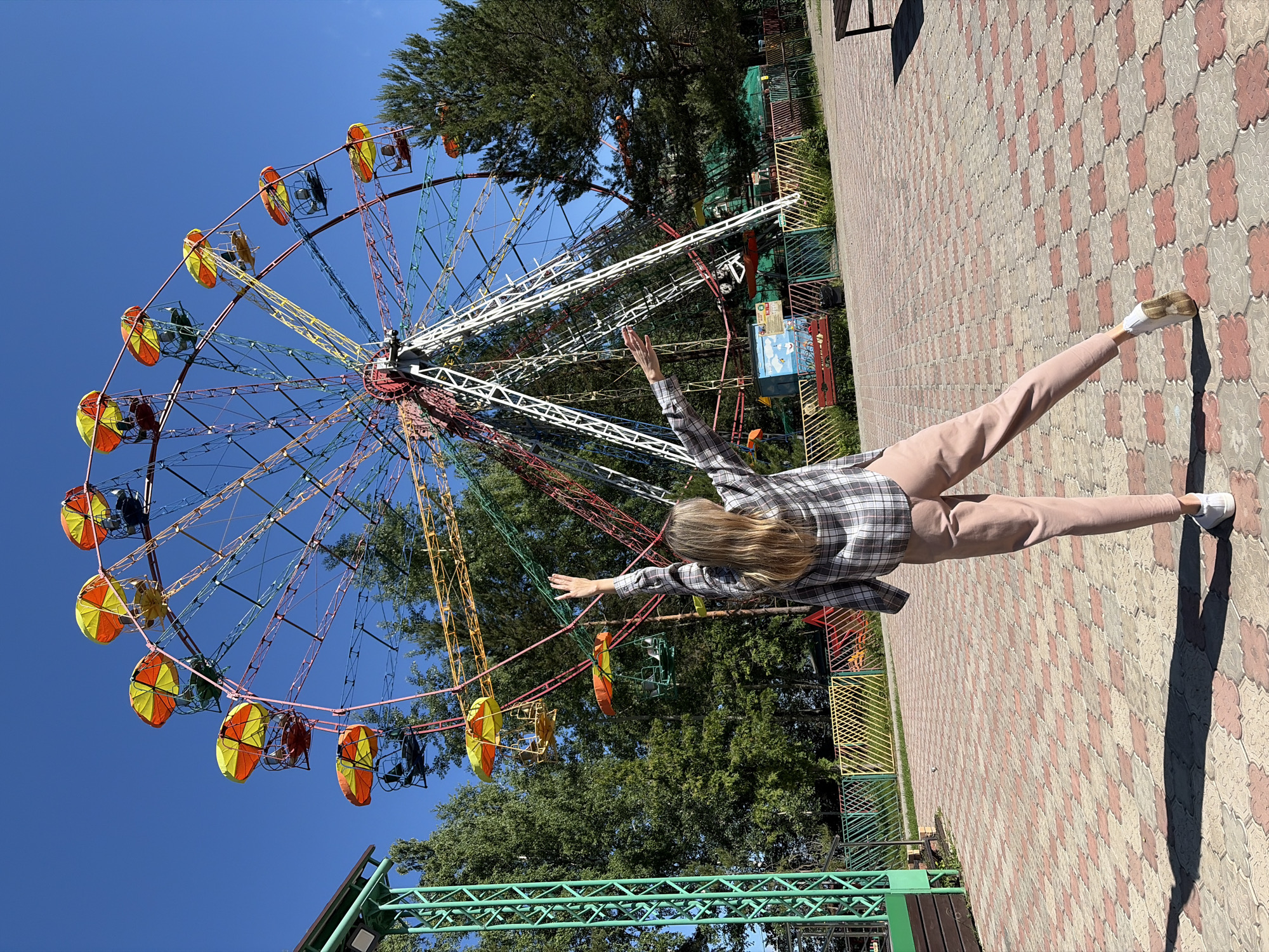 A young woman with long blonde hair is standing in an amusement park in Chaikovsky, Russia, with her back to the camera, facing a colorful Ferris wheel under a clear blue sky. She is wearing a plaid shirt and pink pants, with her arms raised as if to embrace the Ferris wheel. The Ferris wheel has various colored cars with yellow and orange umbrella-like canopies, and a white structural support. In the background, there are green trees, a colorful fence, and paved ground with a reddish-brown and grey patterned brickwork. To the left, a portion of a green metal structure is visible. The scene captures a bright, sunny day at the amusement park.