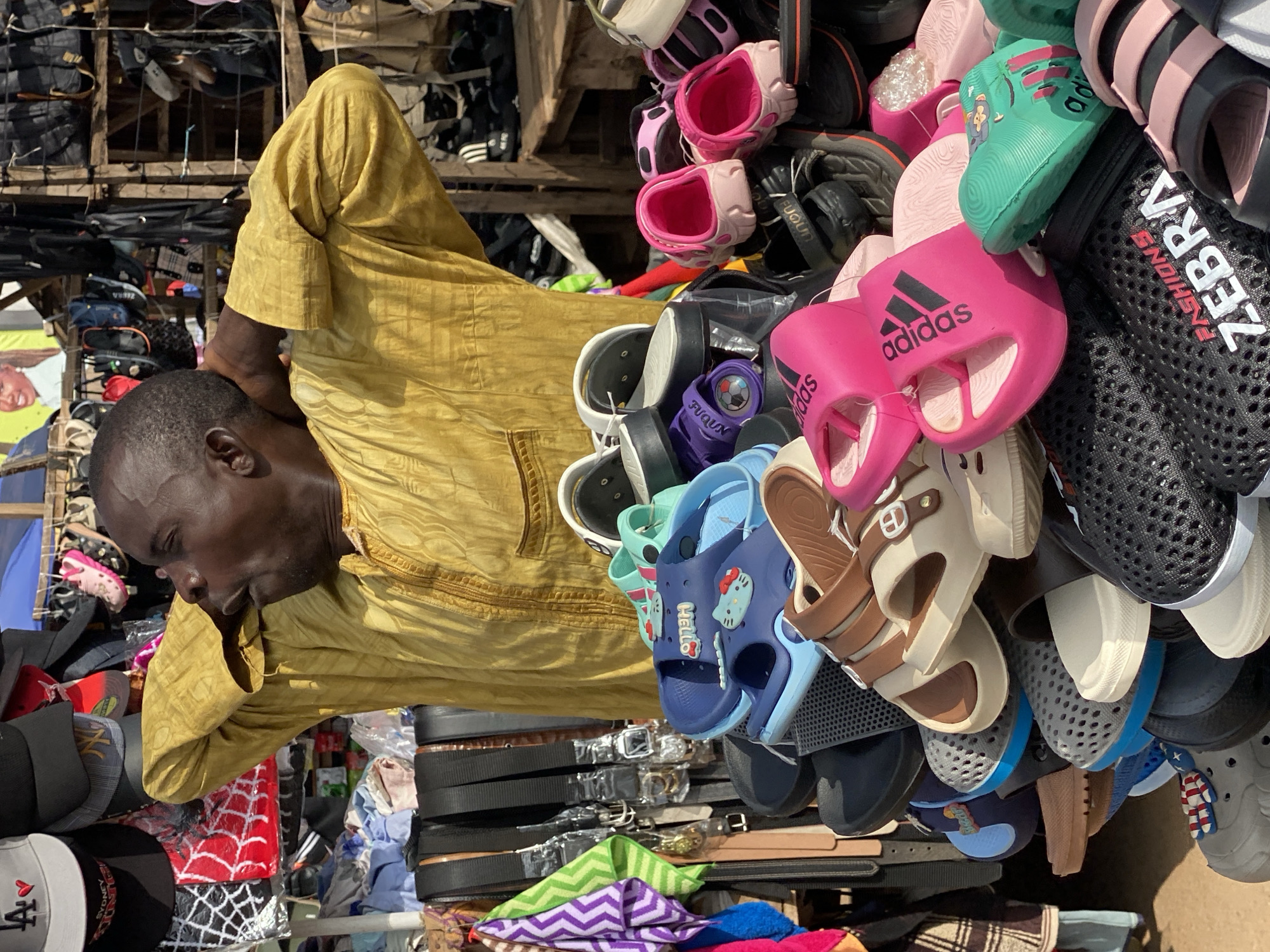 A man in a yellow robe stands with his arms behind his head, his mouth open as if speaking or shouting. He is positioned behind a stall filled with an assortment of footwear, predominantly sandals and flip-flops, in various colors and styles. The stall also displays belts and hats in the background. The scene appears to be an outdoor market in Sabon Tasha, Nigeria, with other market stalls visible in the blurred background. The lighting suggests it is daytime.
