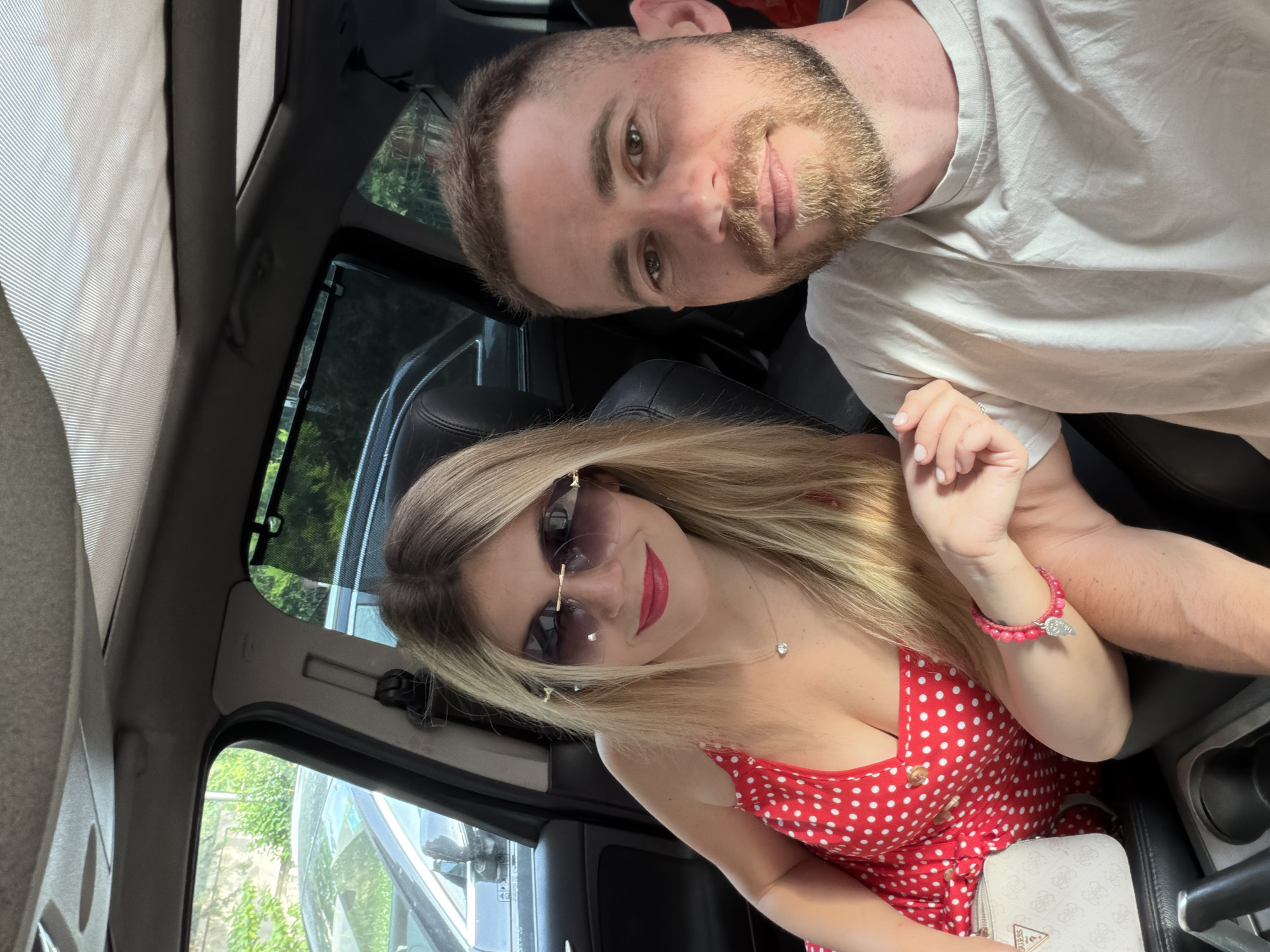 A couple is sitting inside a car. The man is wearing a white t-shirt. The woman is wearing a red polka-dot dress and sunglasses, and she is holding a white Guess purse. They are likely in Budva, Montenegro, though visual confirmation is not present in the image. The car appears to be parked, with a view of greenery and another vehicle through the windows.