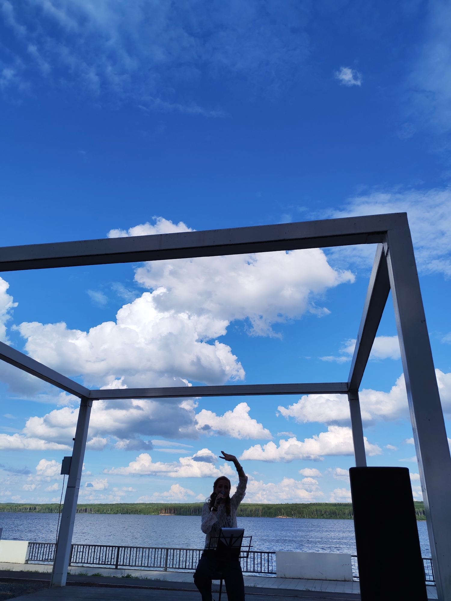 A woman is singing with a microphone while standing on a stage. She is wearing a white lace top and dark pants, and her right arm is raised in the air. The stage appears to be outdoors, under a light-colored canopy structure that frames the sky. The background features a large body of water, likely a river or lake, with a treeline on the opposite shore. A black speaker is visible on the right side of the stage. The sky is a vibrant blue with fluffy white clouds, suggesting it is daytime and the weather is fair. The scene has a relaxed, perhaps performative atmosphere, with the singer engaging with the audience. The location is identified as Chaikovsky, Russia. No visible text is present in the image.