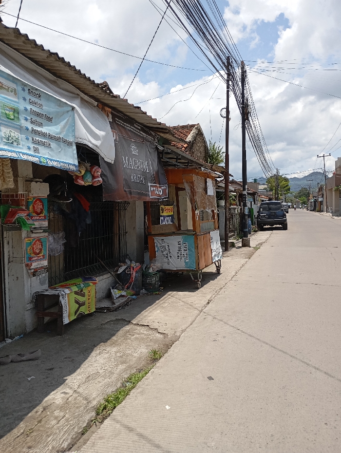 The scene is a street in Katapang, Indonesia, located at latitude -6.998 and longitude 107.552. The street is paved and lined with buildings on both sides. There are numerous power lines running overhead. On the left side of the street, a small wooden cart with a sign that says "MAGNUM KRITE" is parked near a building with a sign advertising snacks, cookies, frozen food, soap, household goods, and cosmetics. In front of the cart, there is a pile of discarded items, including a black bucket, a green bucket, a white bag, and a green bag. There is also a wooden bench with a white and yellow bag resting on it. On the right side of the street, a black car is parked on the shoulder of the road. The street is empty, except for the car. The sky is partly cloudy. The buildings are painted in muted colors and appear to be in disrepair. The overall atmosphere is quiet and peaceful.