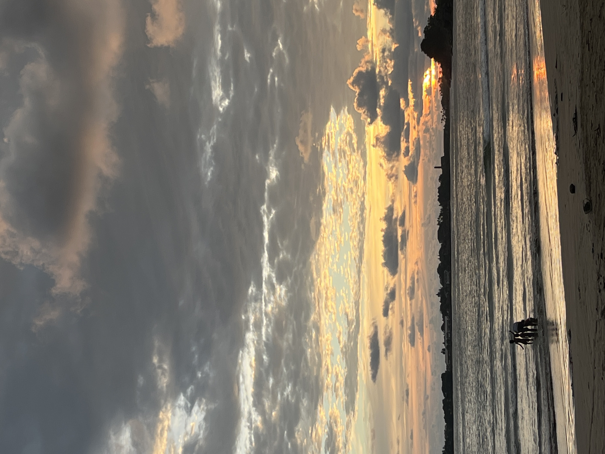 The image depicts a sunset at a beach in Galle, Sri Lanka. The sky is filled with dark, heavy clouds on the left side, transitioning to lighter clouds illuminated by the setting sun on the right. The sun is partially obscured by a treeline, with a tall structure visible in the distance on the horizon. The ocean reflects the light of the sunset, creating a shimmering effect on the water's surface. Several figures, possibly people, are standing in the shallow water near the beach. The beach sand is visible in the foreground.