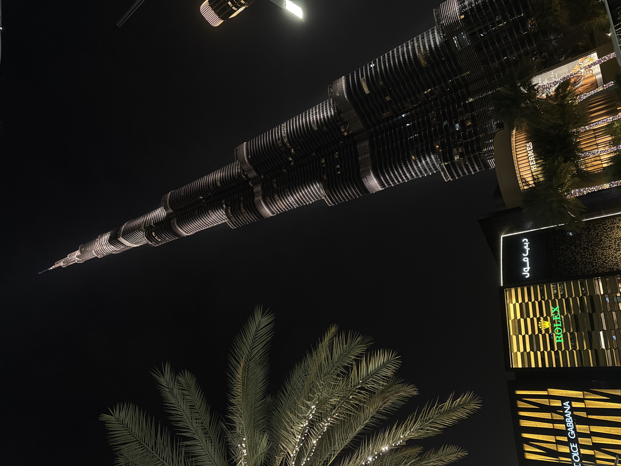 The image, taken at night in Dubai, United Arab Emirates, features the Burj Khalifa illuminated against the dark sky. Palm trees with lights are in the foreground. Adjacent to the tower are high-end retail stores with prominent signage including Rolex and Dolce & Gabbana. The Dubai Mall sign is also visible.