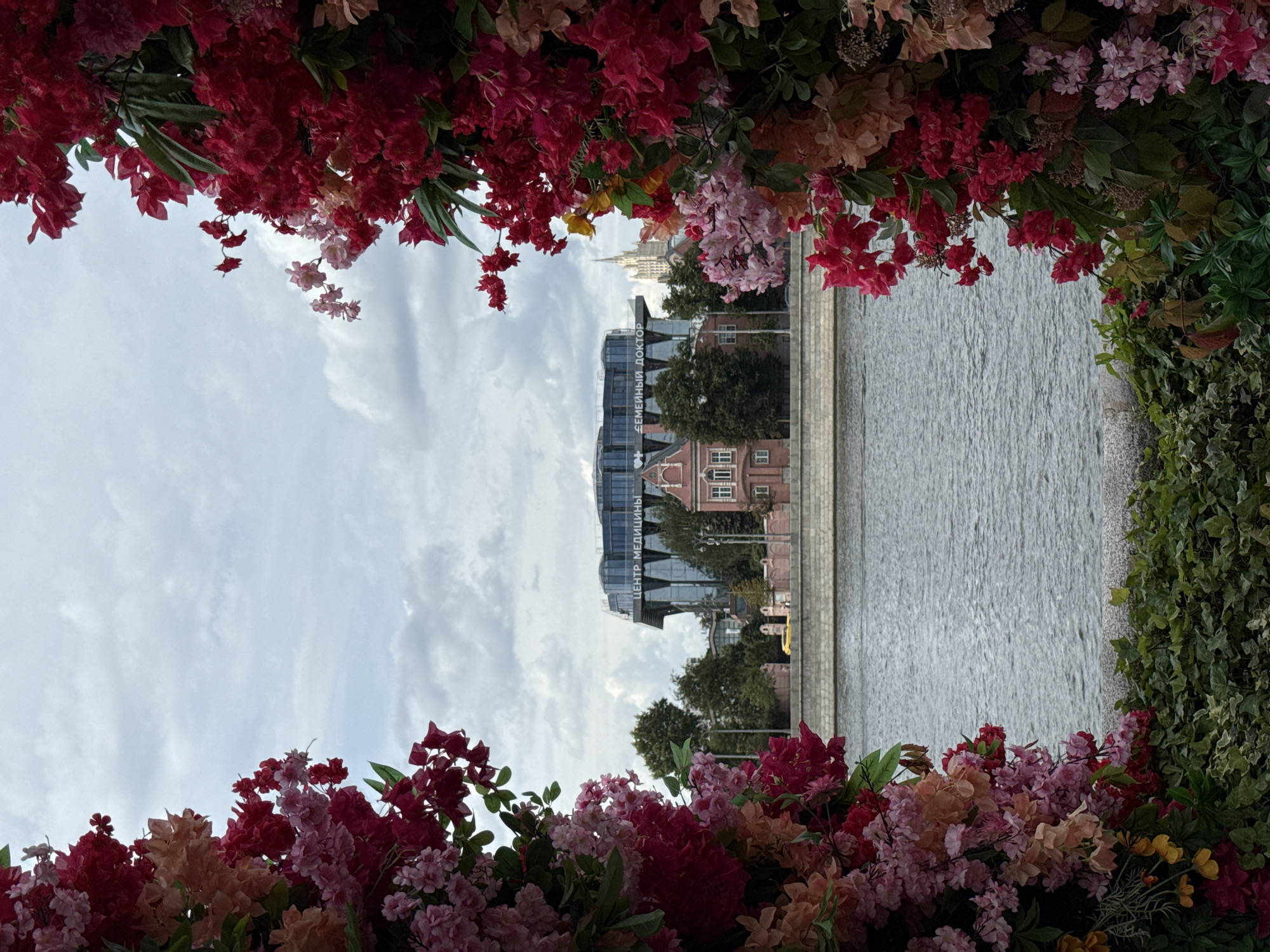 An outdoor, eye-level, medium shot captures a floral archway framing a view of a river, buildings, and the sky. The archway is densely packed with artificial flowers in shades of red, pink, and peach, with lush green foliage. The river flows in the foreground, separated from the bank by a stone embankment. Across the river, a row of trees stands behind a stone wall. Beyond the trees, several buildings are visible: a small, traditional pink building with white trim and a larger, modern glass building with a curved roof. Text on the glass building reads "ЦЕНТР МЕДИЦИНЫ" and "СЕМЕЙНЫЙ ДОКТОР". A tall, spire-like building is visible in the distance. The sky is overcast with scattered clouds.