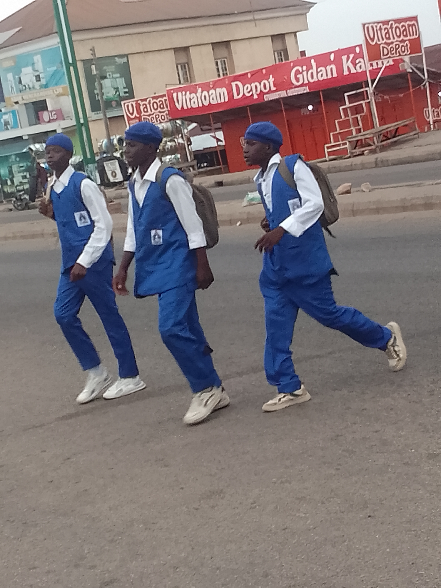 In Pantame, Nigeria, three young men in matching blue uniforms and berets are walking across a street. They appear to be students, each wearing a white shirt and blue vest with an emblem. Two of the men have backpacks slung over their shoulders. They are walking on a paved road in front of a row of red buildings, one of which displays a "Vitafoam Depot" sign. The background shows other buildings and some vegetation, suggesting an urban setting. The lighting indicates it is daytime.