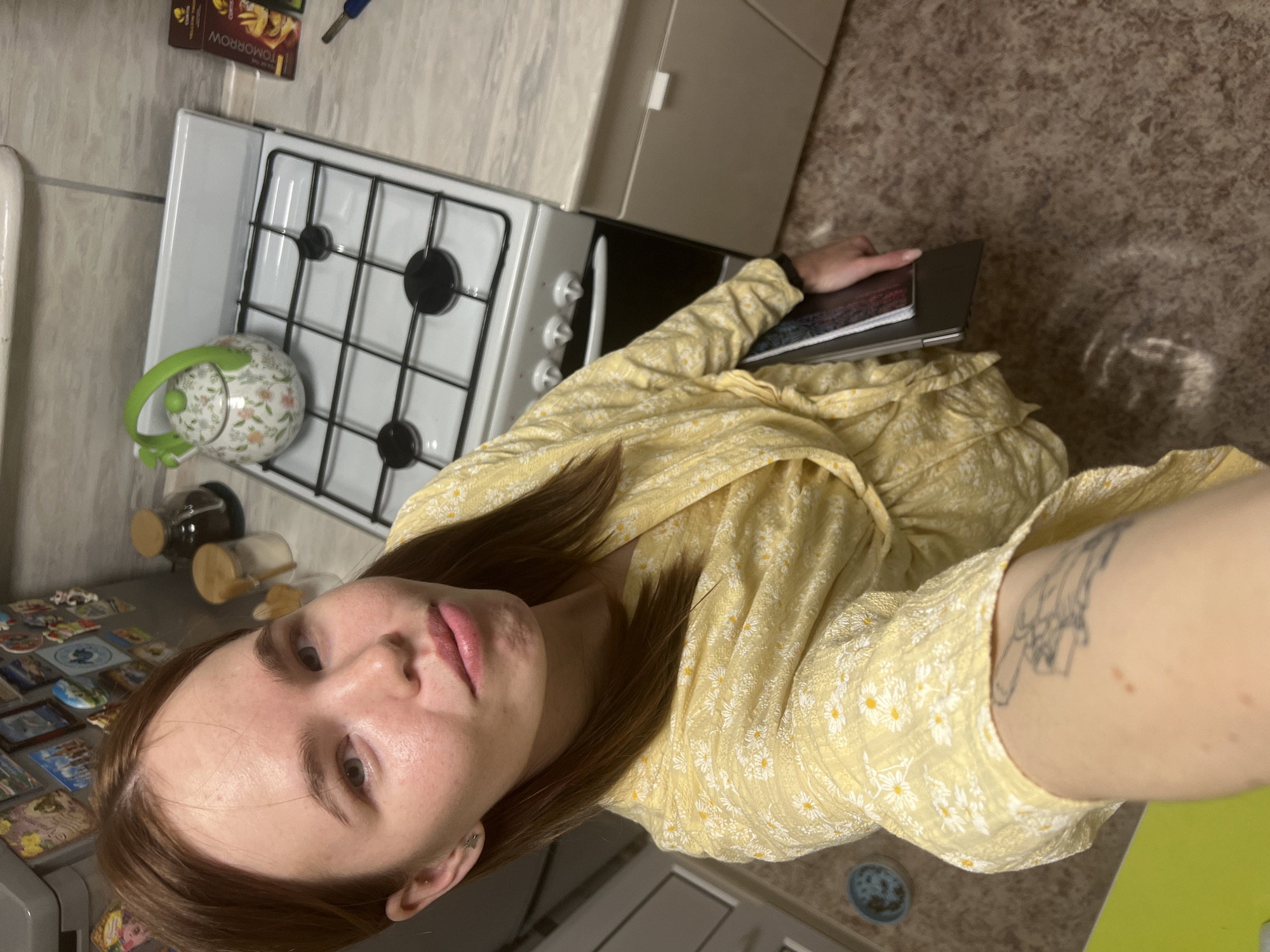 A woman is taking a selfie in a kitchen. She has brown hair and is wearing a yellow dress with a floral pattern. Her left arm, visible in the lower right of the frame, has a tattoo. She is seated and has a laptop and notebook on her lap. Behind her is a kitchen counter with a stove, a kettle, and canisters. There are boxes of "TOMORROW" brand cookies visible near the stove. The floor has a brown speckled pattern. The location is in a kitchen in Chaikovsky, Russia.