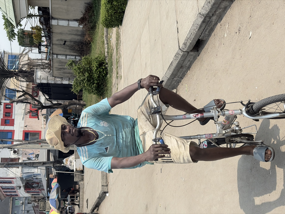 A dark-skinned adult male is shown riding a silver-colored folding bicycle on an unpaved road in Jakande, Nigeria. He wears a straw hat, a light blue short-sleeved Adidas Real Madrid t-shirt, light-colored (possibly denim) shorts, and grey slide sandals. His face features a short dark beard, and he has black earphones/earbuds around his neck. He wears a gold-colored chain necklace, a white beaded rosary-style necklace, and a dark wristwatch on his left wrist. His hands grip the bicycle's handlebars, and his gaze is directed forward with a neutral expression. A sticker depicting "Jesus" is visible on the bicycle's frame. To the right, a concrete curb and sidewalk border the road. The background reveals an urban environment with multi-story buildings, including one with prominent red and white sections and blue-framed windows, and another with a dark metal gate. Multiple utility poles with numerous wires are present, alongside sparse vegetation like bushes and a palm tree. On the far left, colorful market umbrellas and a white container are visible, indicating a commercial area. The scene is brightly lit by daylight, casting shadows.