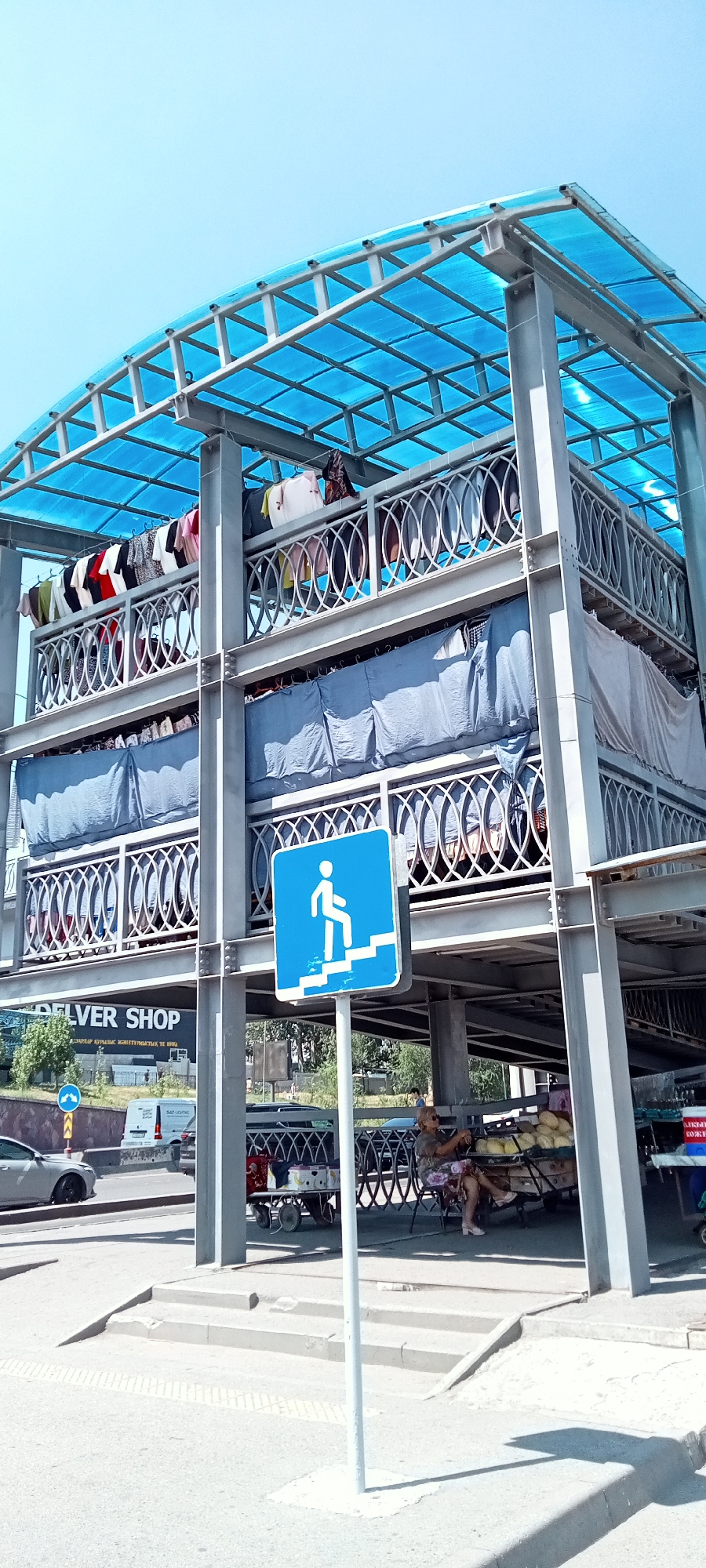 The image captures an outdoor market scene under a blue, translucent roof supported by a grey metal frame structure. The structure appears to be multi-leveled, with laundry hanging on clotheslines on the upper levels. A blue pedestrian sign with white stairs and a person climbing is prominently displayed on a pole in the foreground.
Below the structure, a woman is seated on a chair next to a cart laden with fruits, possibly melons. Other market stalls and carts are visible in the background, with a white van parked nearby. A silver car is seen on the road in the left of the image. The text "DELIVER SHOP" is visible on a sign on a building in the background, suggesting a commercial area. The environment is a clear, sunny day.
Based on the presence of the "DELIVER SHOP" sign and the architectural style, the location is likely a marketplace or a commercial district in Almaty, Kazakhstan. The scene depicts a typical day in a bustling urban market, with everyday activities of people selling goods and shoppers browsing.